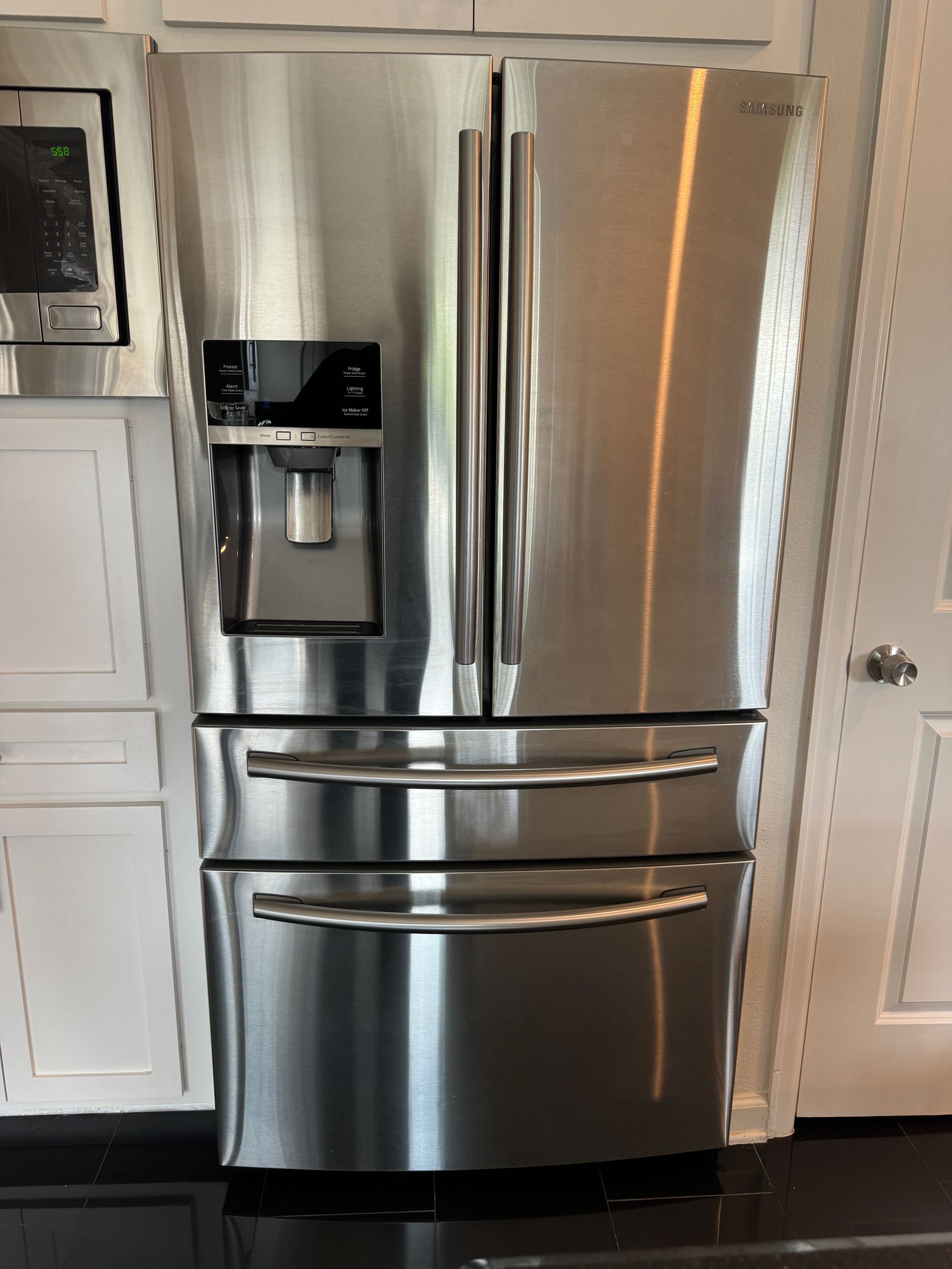 Stainless steel French door refrigerator with a water dispenser, set in a kitchen with white cabinets.