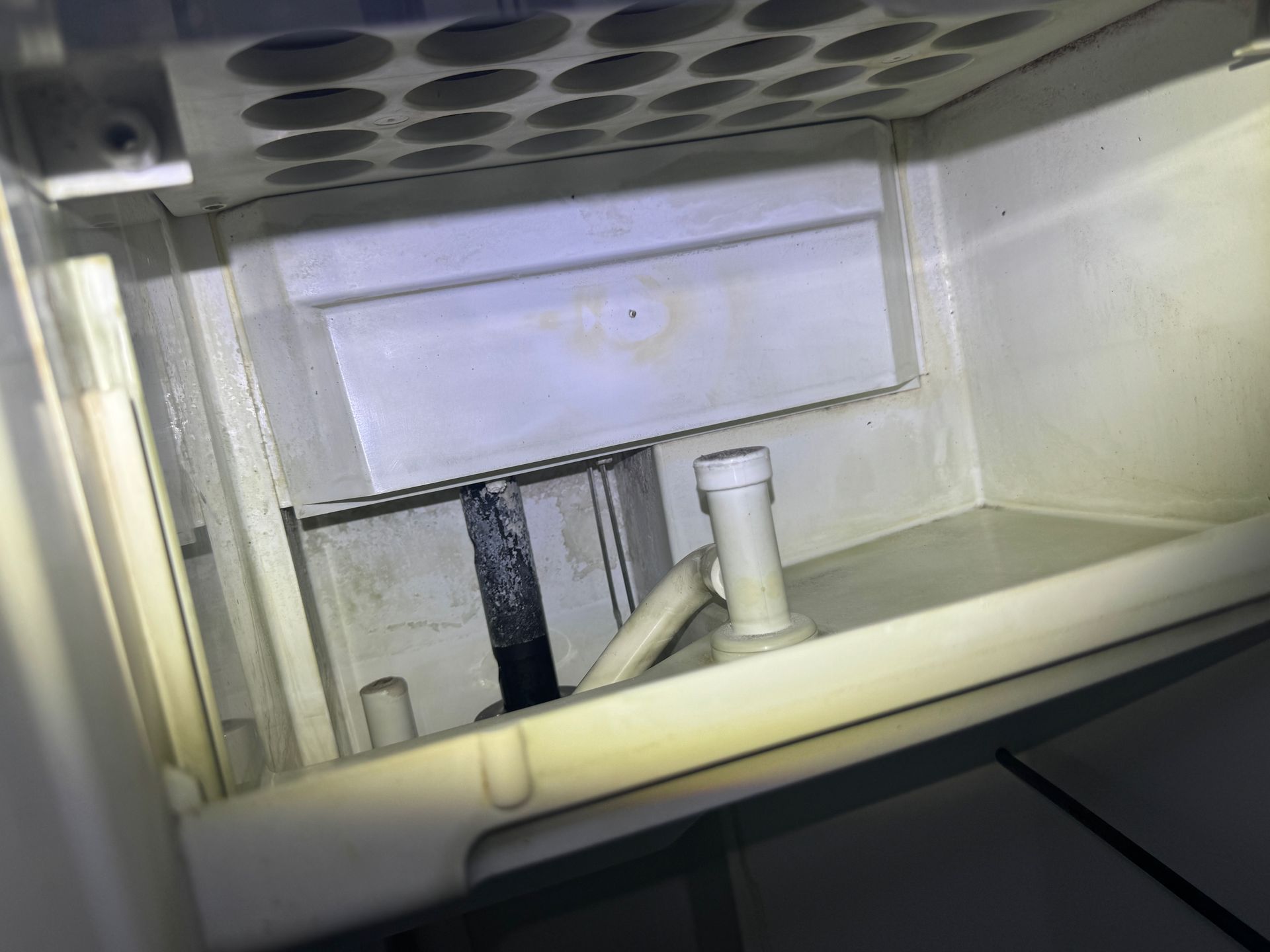 Interior of an ice maker compartment, showing a white plastic basin with a drain pipe, a black, vertical component, and a partially visible grid above.
