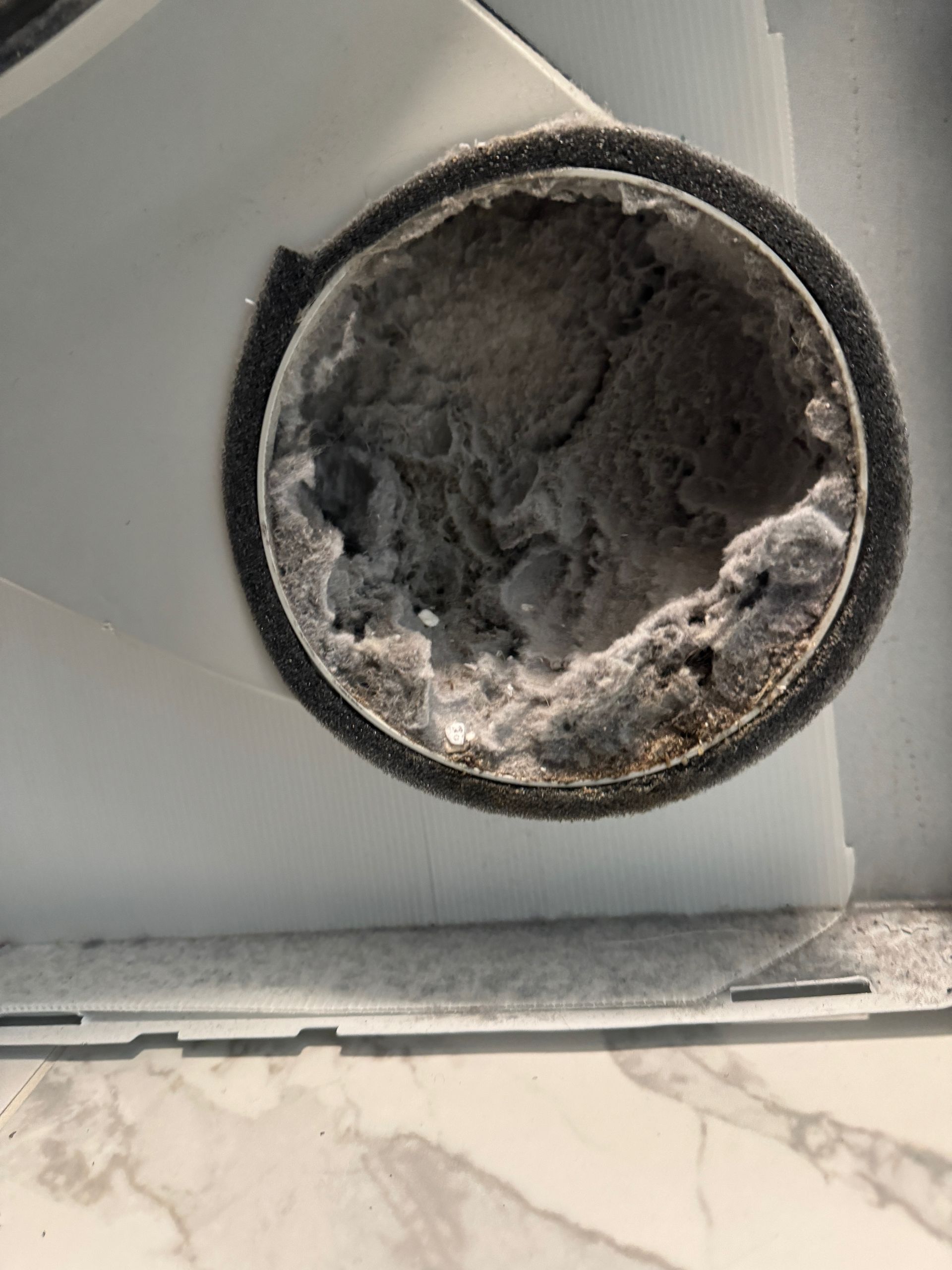 Close-up of a dryer lint trap, clogged with thick gray lint. The opening has a dark, dusty appearance.