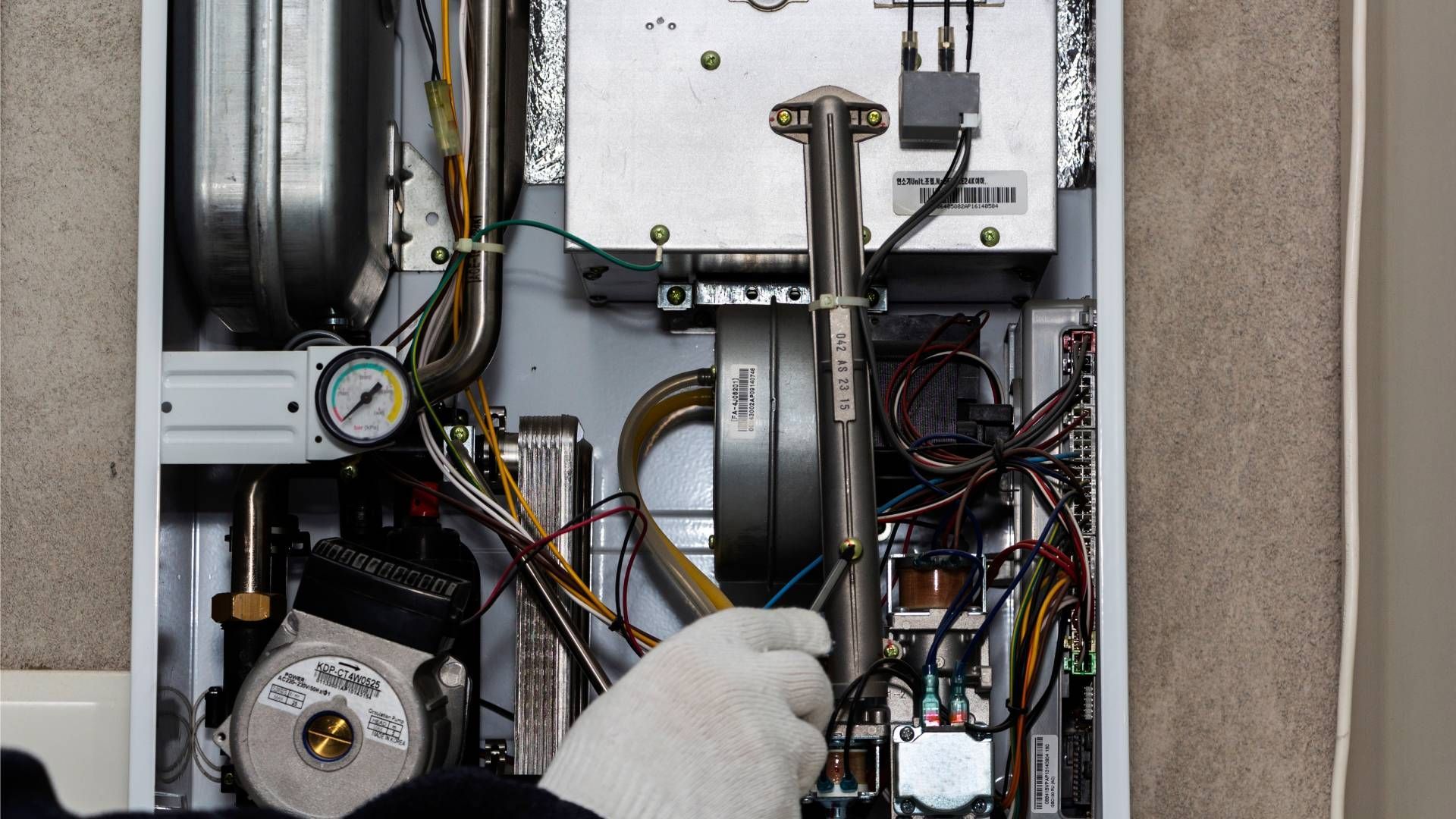 A person in gloves working on the inside of a furnace or boiler. Wires, pipes, and components are visible.