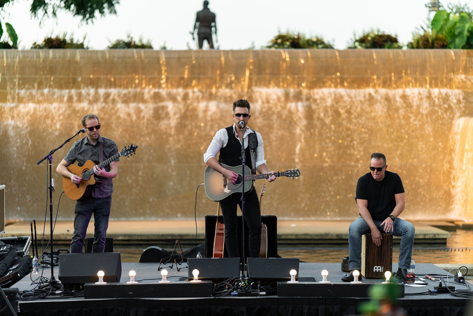 Three men are playing guitars and singing on a stage in front of a waterfall.
