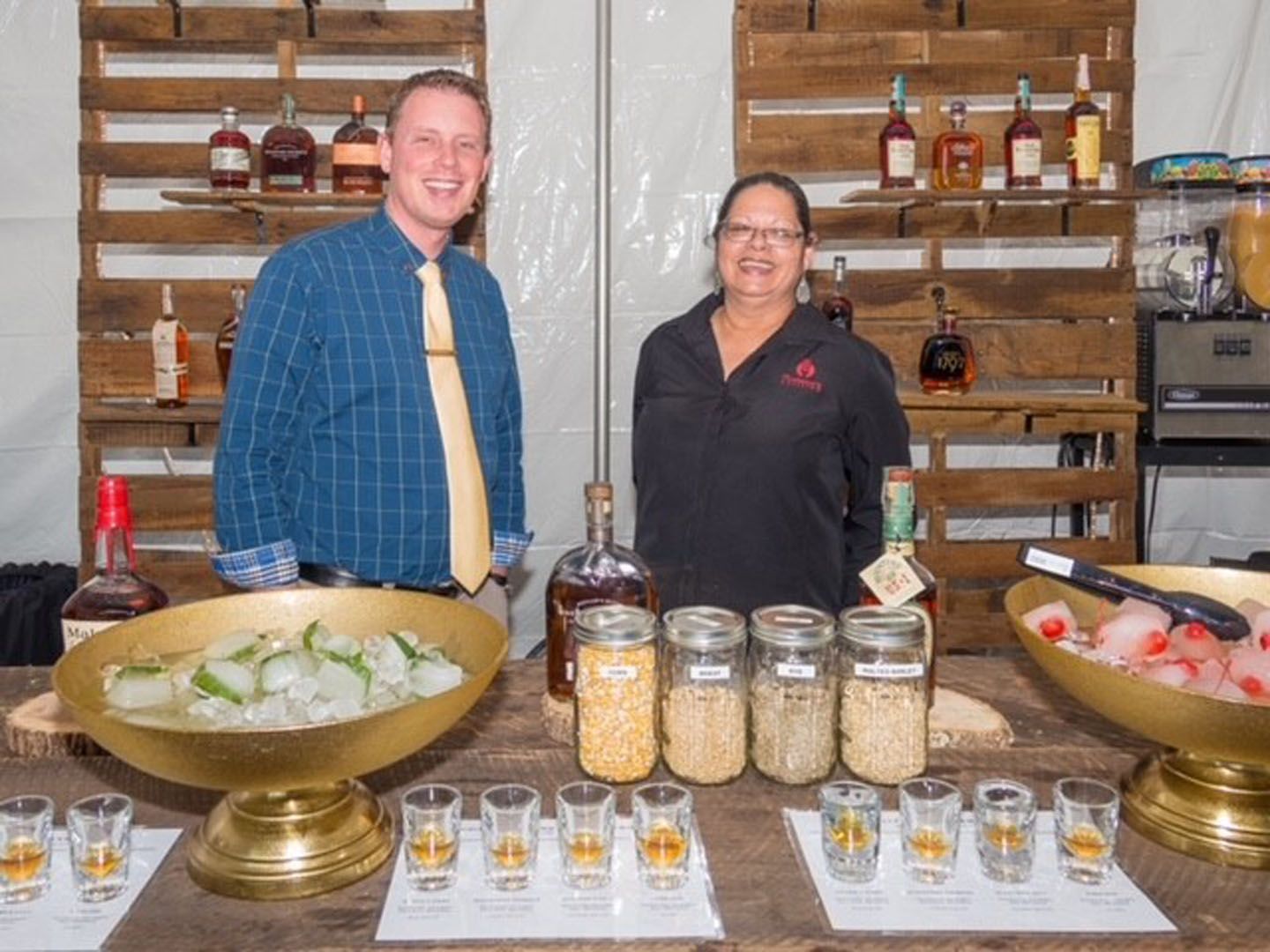 A man and a woman are standing behind a bar filled with bottles of alcohol.