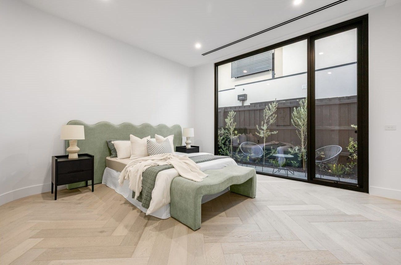 Bedroom with light wood floor, a green bed, and a large window overlooking a small garden.