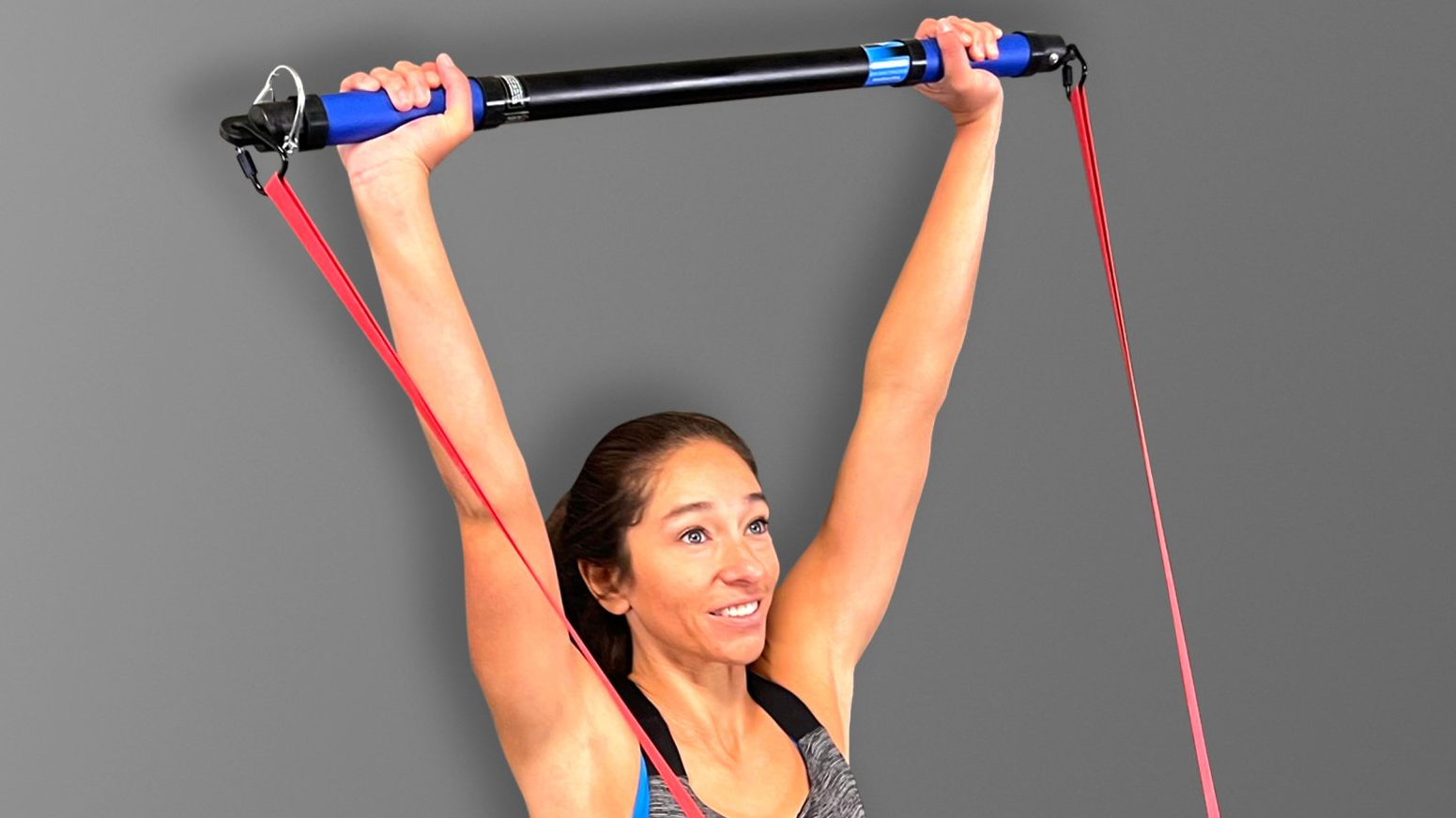 a woman is standing and performing a SmithShaper resistance band shoulder press exercise using an S2-BAR - a 33-inch resistance band bar connected to a red SmithShaper 42-inch Zobo Sports High Stretch Therapy Band that is anchored with a door anchor that is not shown.