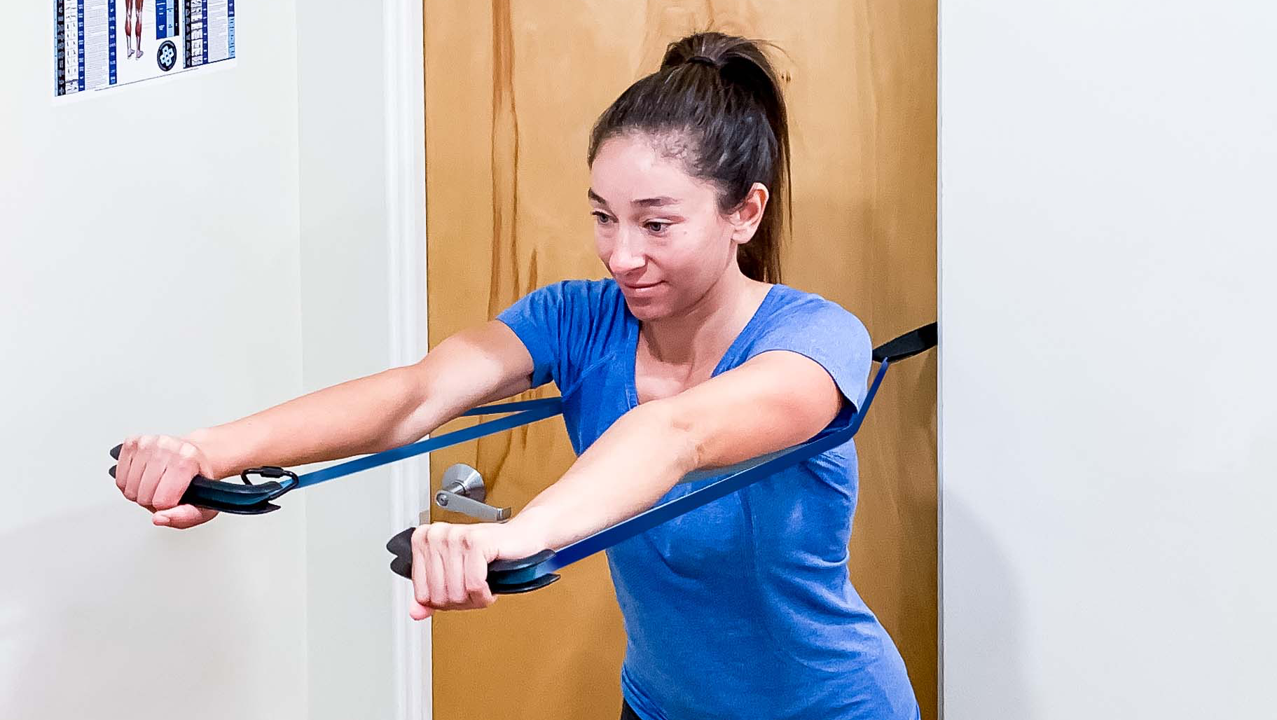 a woman in a blue top is standing and performing the SmithShaper chest press resistance band exercise using SQUATX Loop Max handles wrapped in a blue 32-inch SmithShaper Zobo Sports High Stretch Therapy Band that is anchored to a door behind her