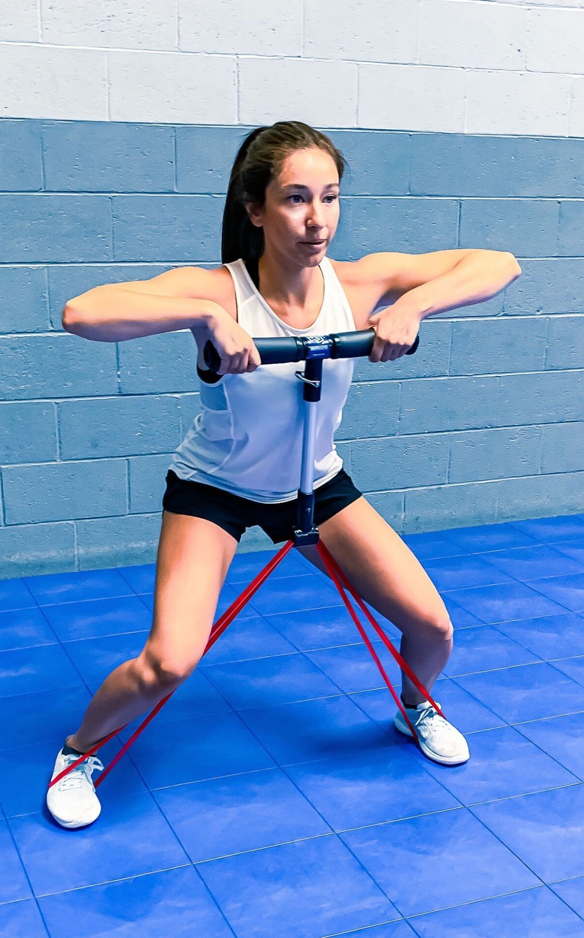 a woman in a white top and black shorts is shuffling as she performs SmithShaper Upright Tow Squat Jacks or Shuffle resistance band exercise using a TR-BAR resistance  exerciser connected to a red SmithShaper 42-inch Zobo Sports High Stretch Therapy Resistance Loop Band anchored on each of her feet