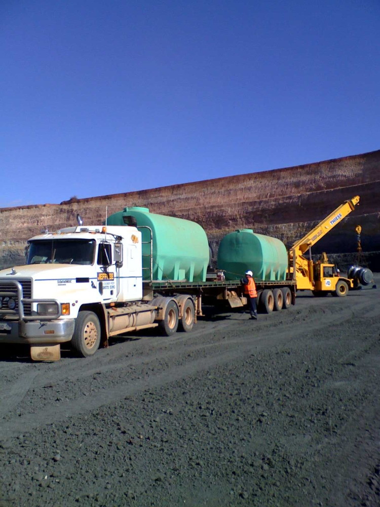 A semi-truck hauling two green water tanks on a flatbed trailer, with a worker and a forklift at a quarry.— Ts Jeffree Contractors in Mount Isa, QLD