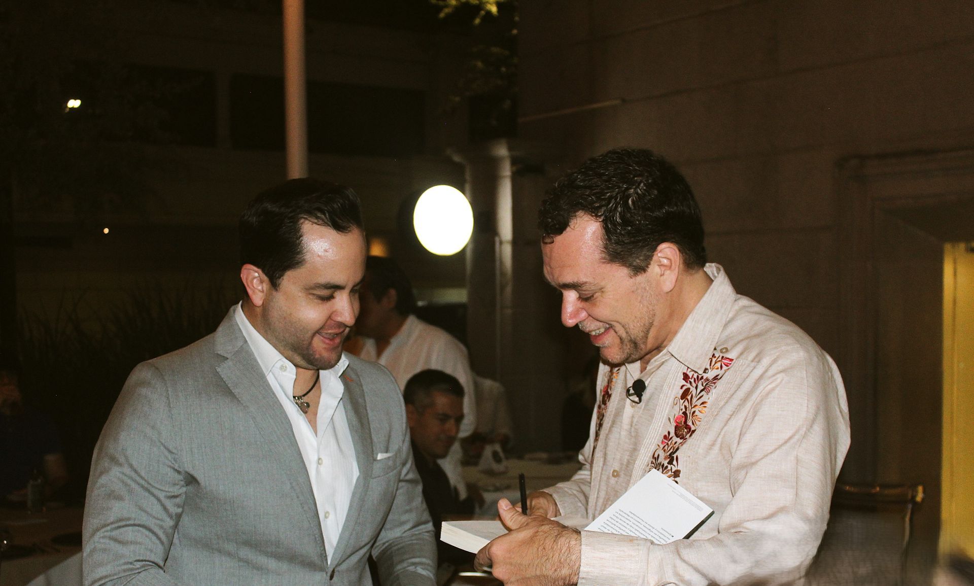 Un hombre firma un libro para otro hombre. En un restaurante. Ambos hombres sonríen.