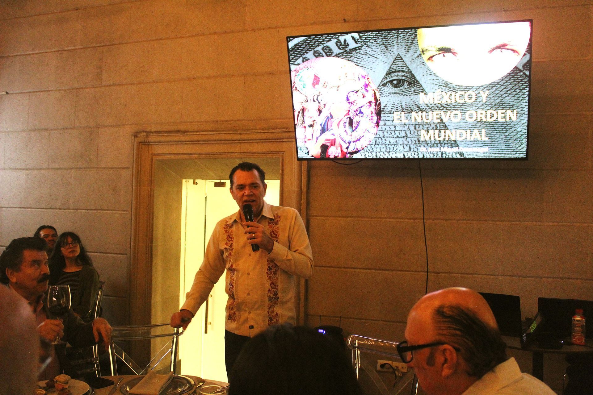 Hombre hablando por un micrófono en un evento. La pantalla muestra el texto: 