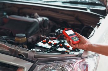 Mechanic testing car battery with a red device in an open engine bay.