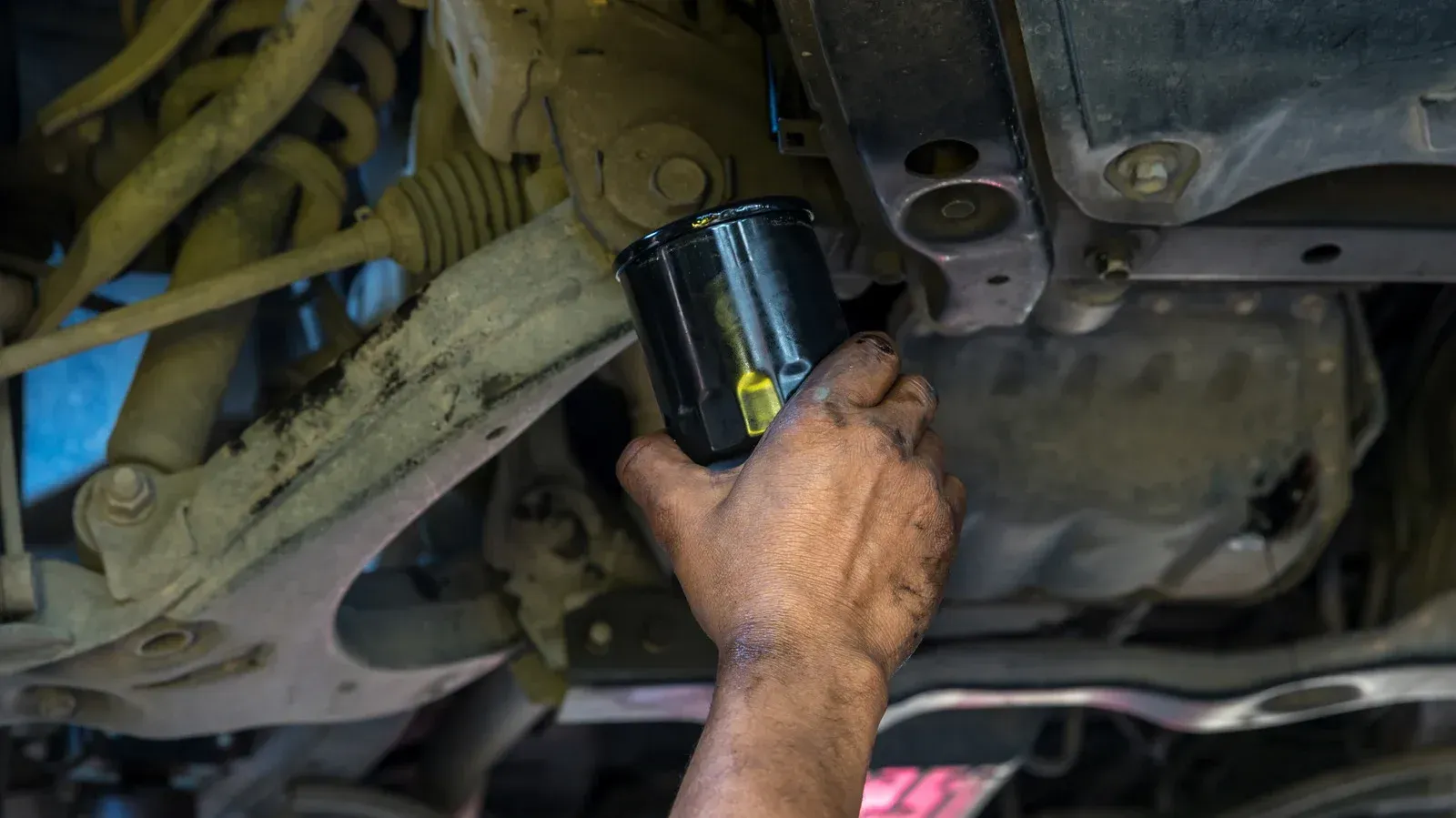 Hand holding a black oil filter under a car, likely during an oil change.