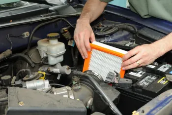 Hands replacing an air filter in a car engine, orange filter, blue car, daytime.