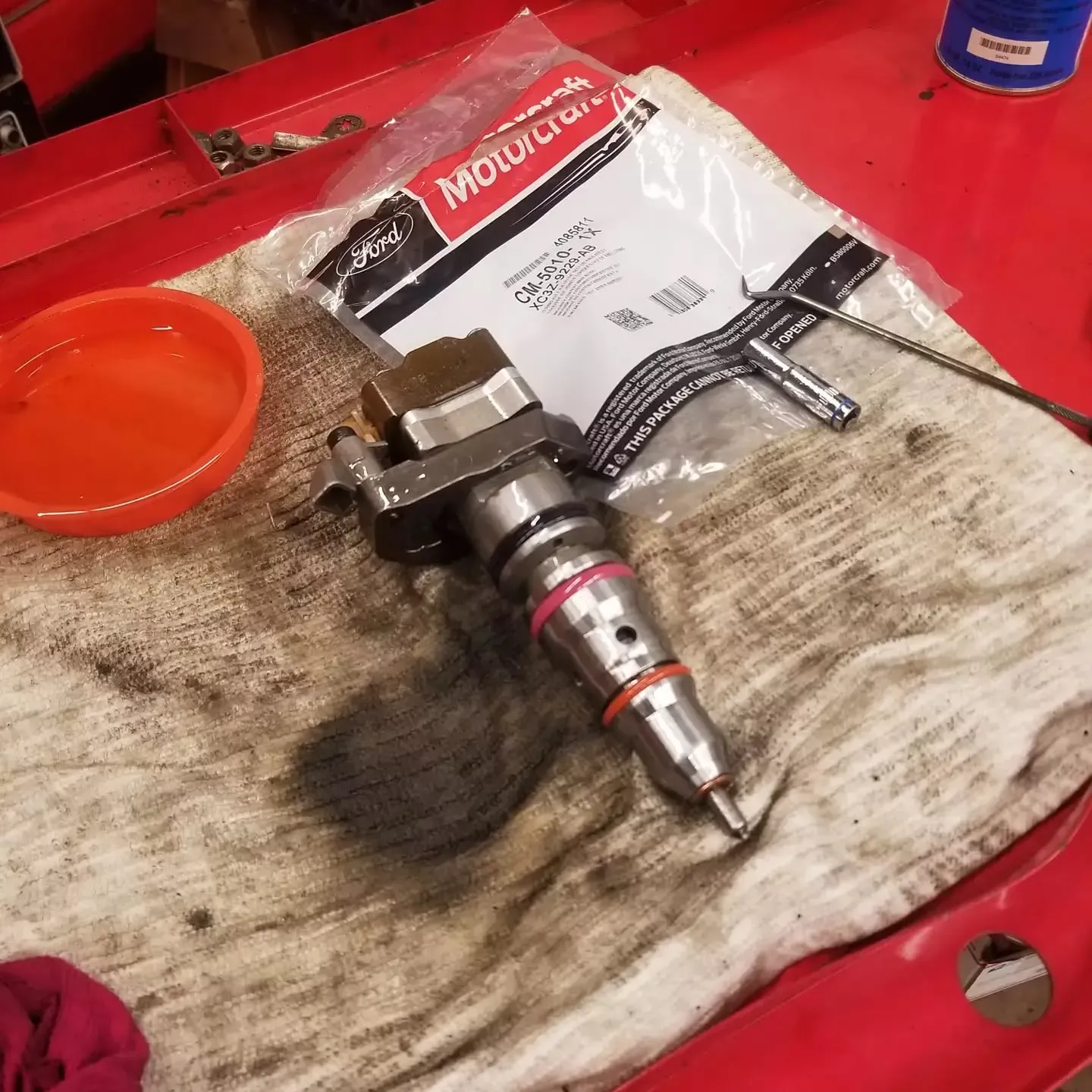 Diesel fuel injector with red o-rings, Motorcraft packaging, orange bowl, and tools on a red surface.