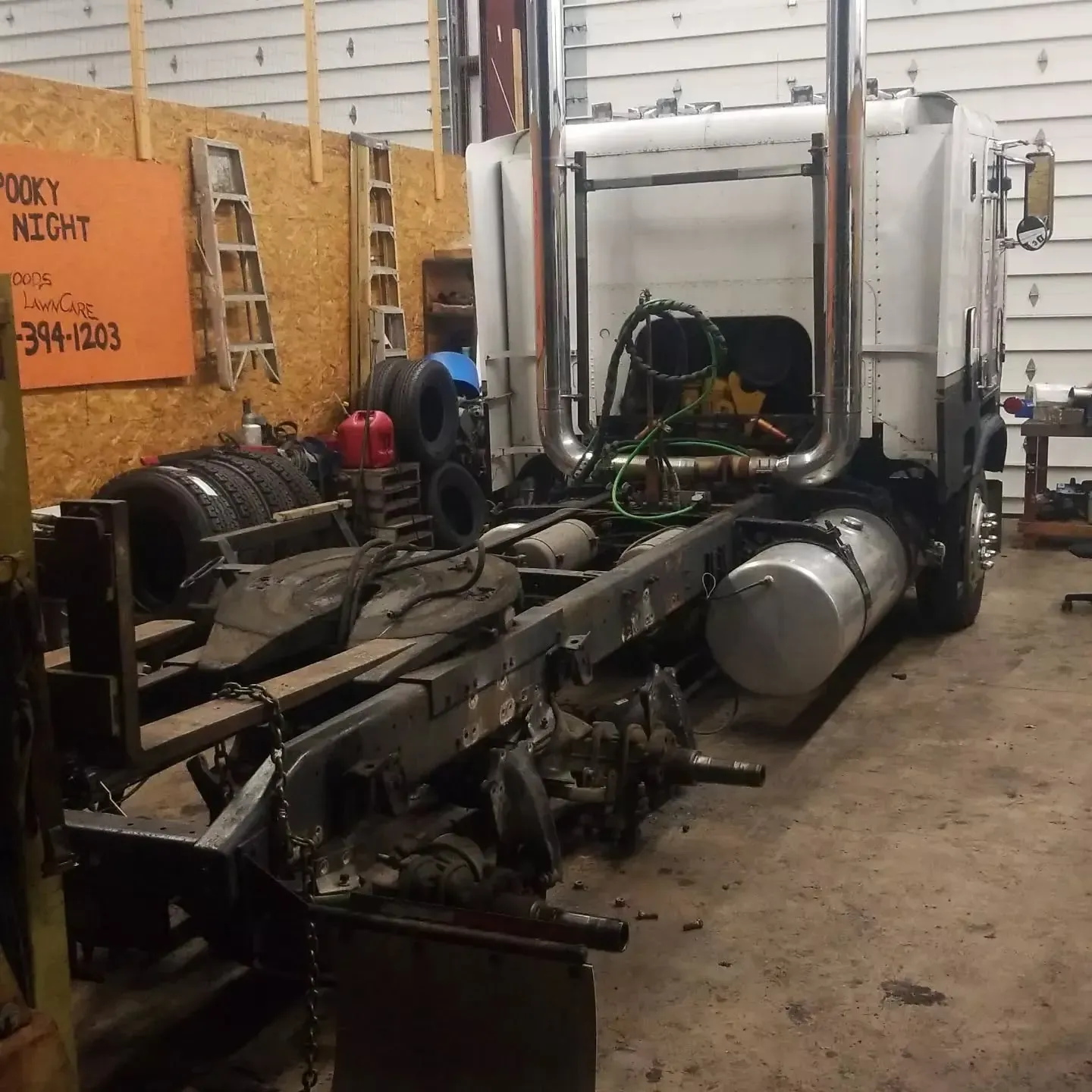 A semi-truck chassis in a garage. The back of the truck is missing its trailer. Tools and tires are visible.