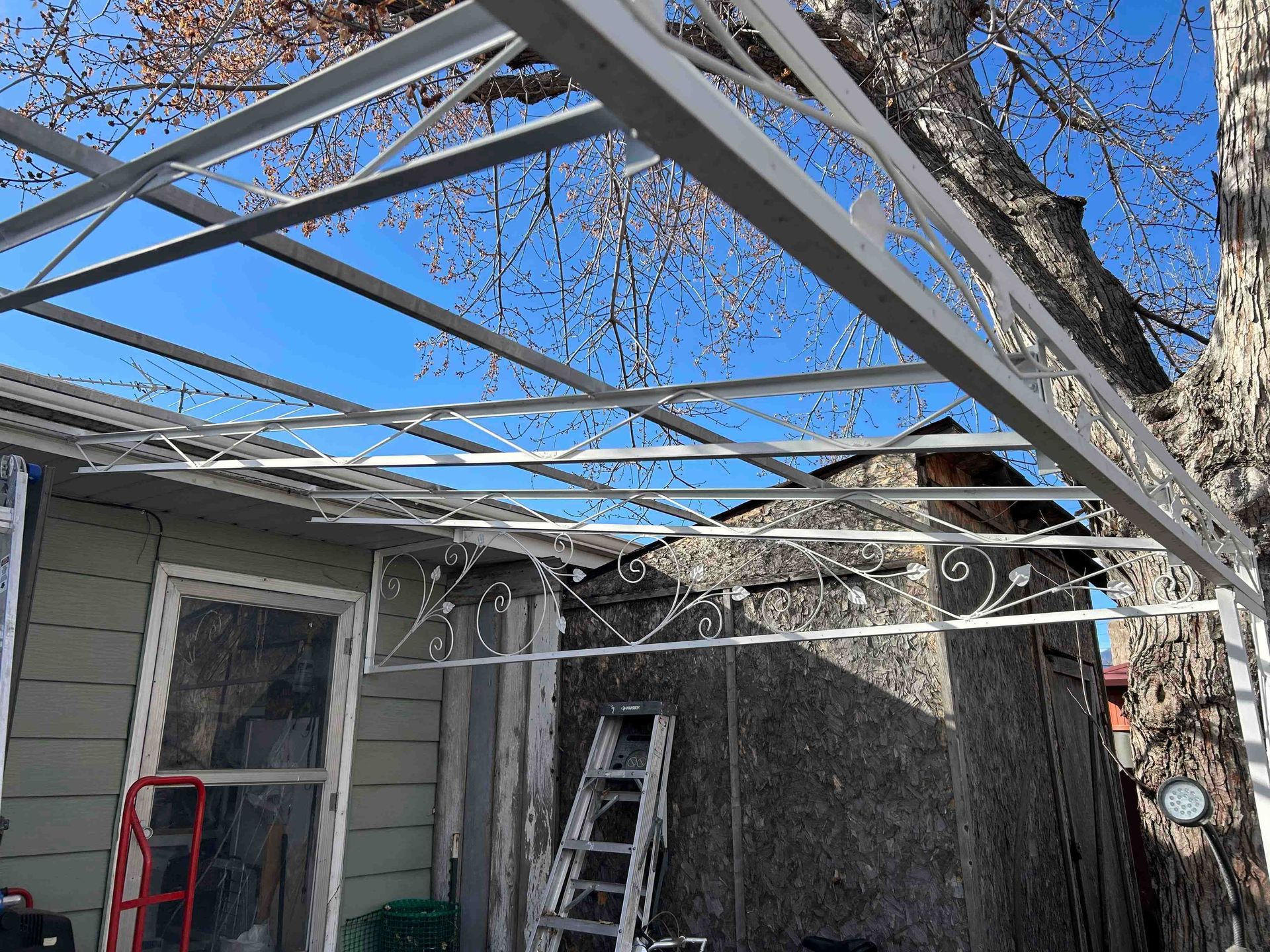 Metal frame canopy extending over a patio, with bare tree branches and clear blue sky in the background.
