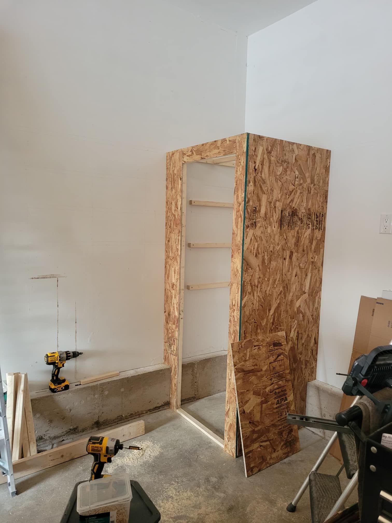 A partially constructed wooden storage cabinet in a garage, featuring OSB siding, and two power drills.