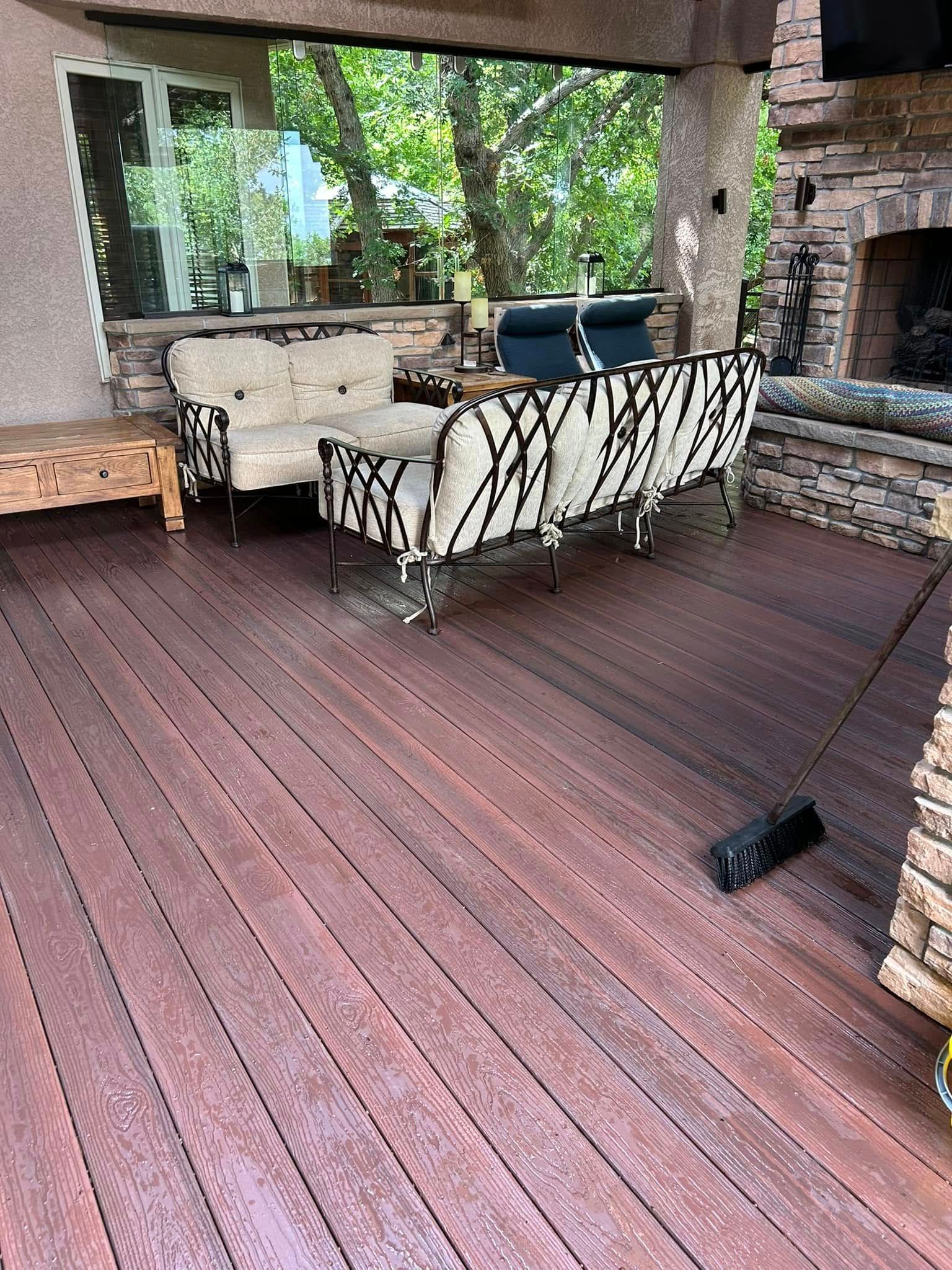 Wooden deck with patterned sofas, fireplace, and lush greenery viewed through a window.