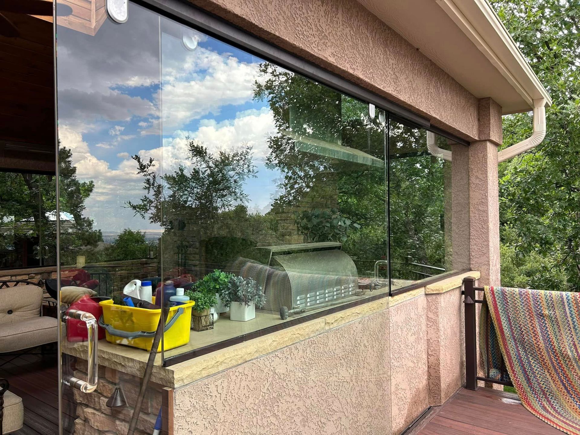 Glass-walled patio with a grill, overlooking trees and a blue sky with clouds, on a sunny day.