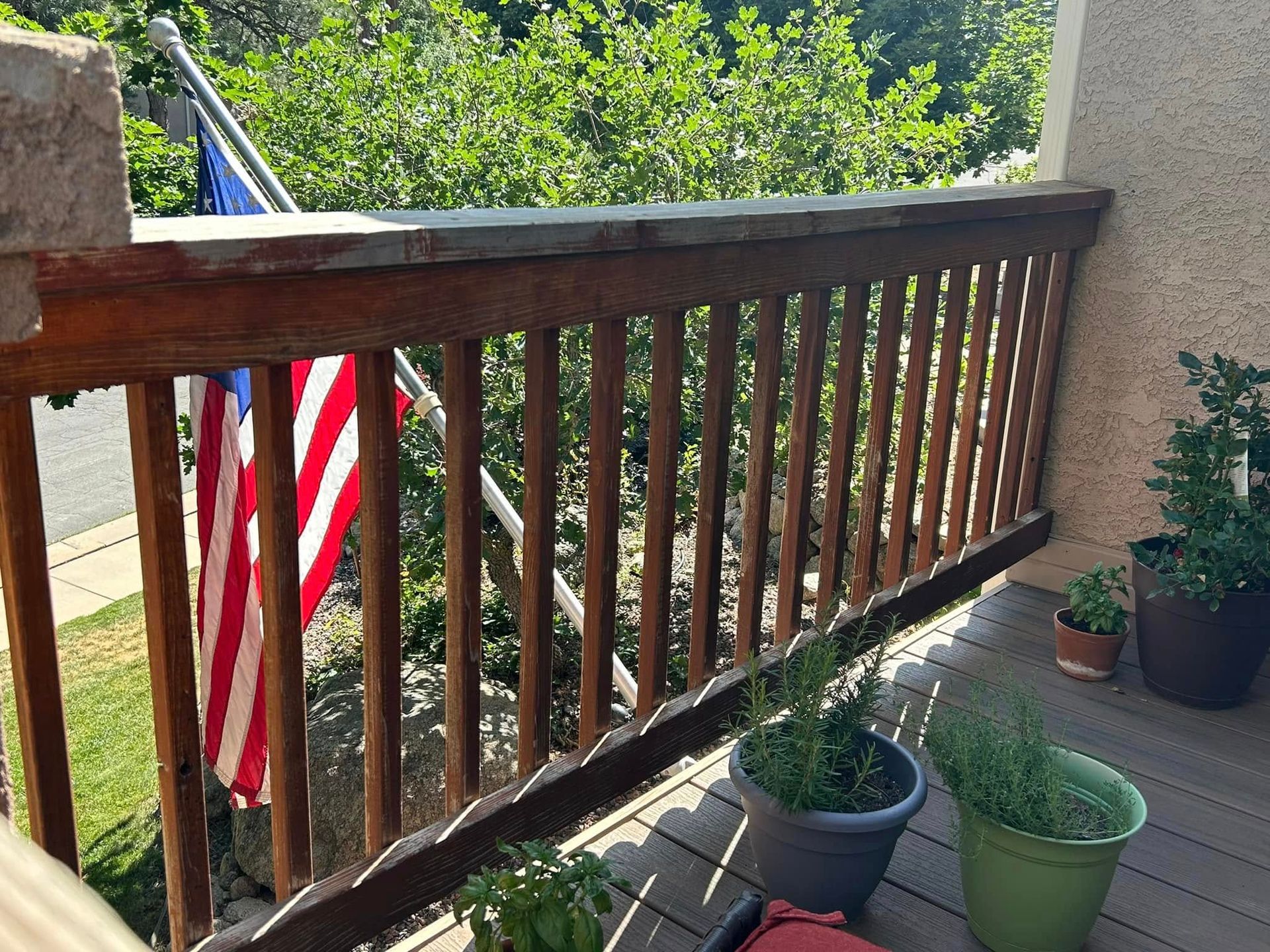 Wooden deck railing with American flag; potted plants and greenery in the background.