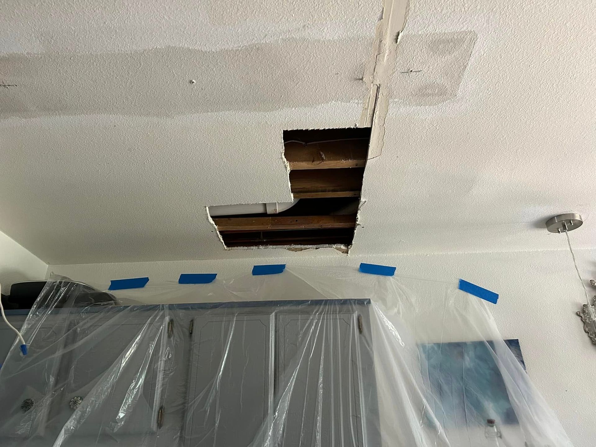 Hole in a textured white ceiling with exposed beams. Blue tape and plastic sheeting cover cabinets below.