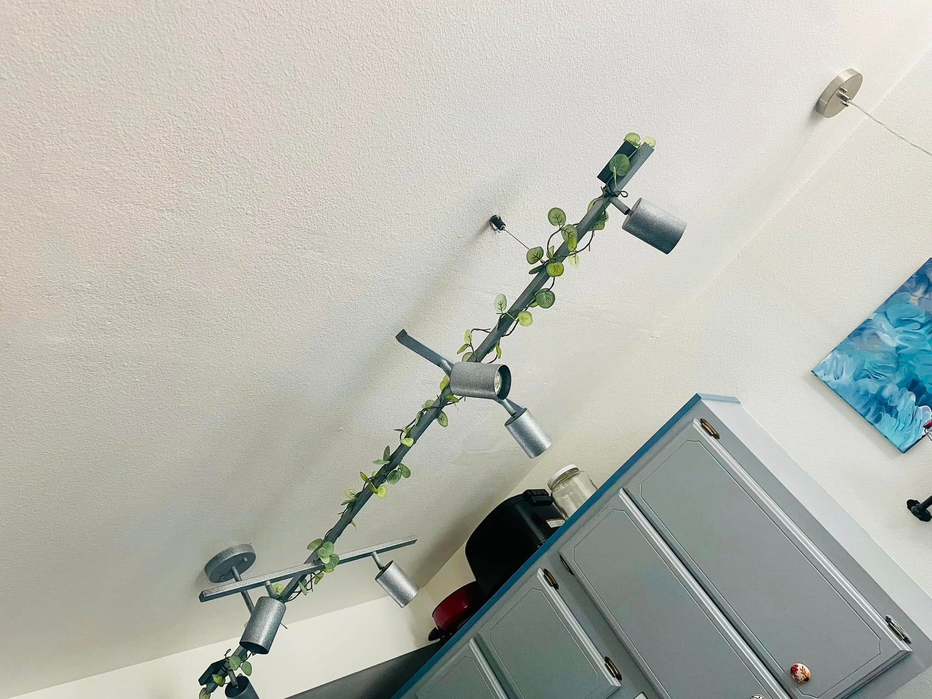 Ceiling-mounted track lighting with spotlights and faux ivy against a white ceiling and gray cabinet.