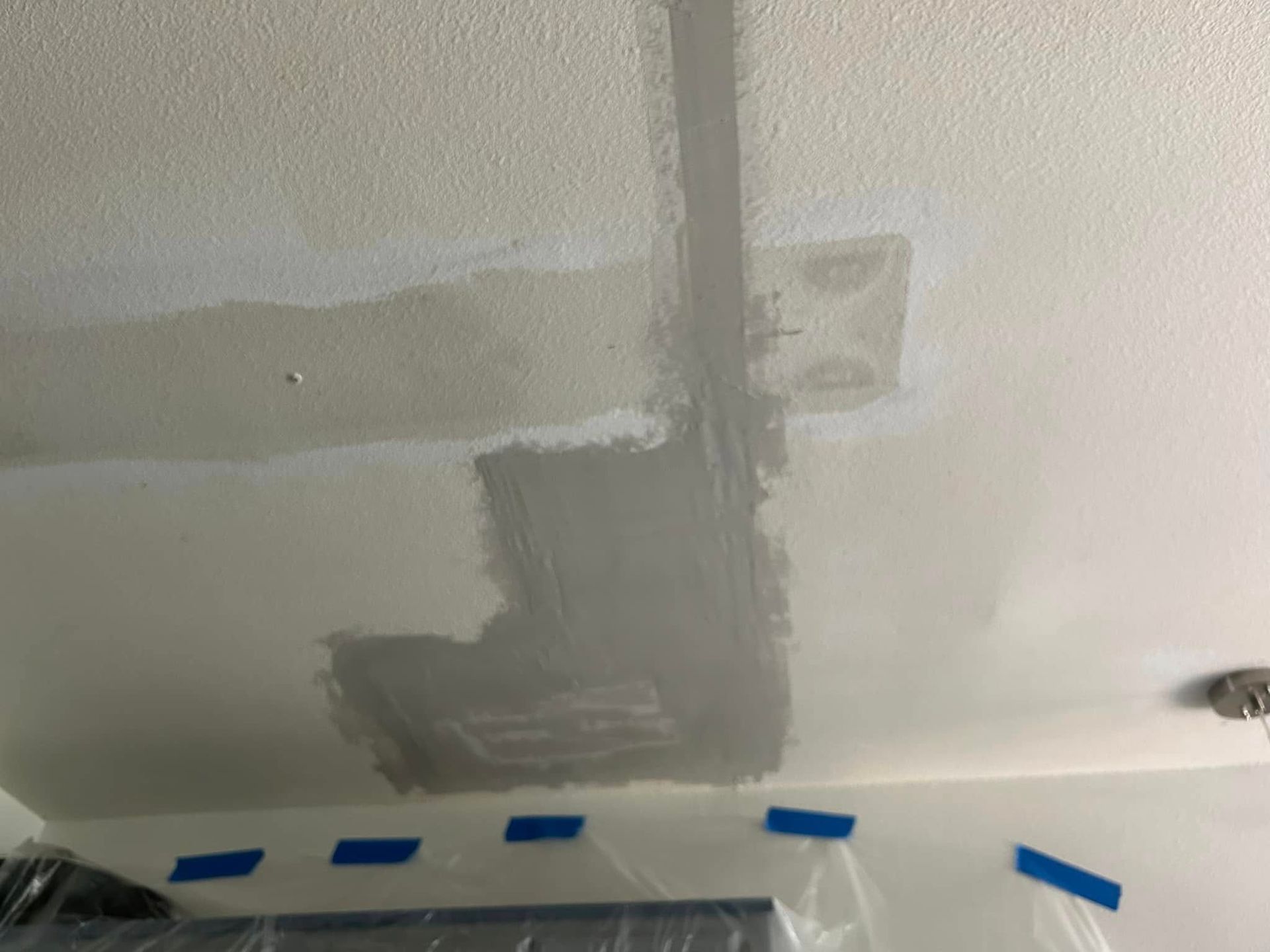 Ceiling with patched areas of gray paint against a white background. Blue tape lines the lower edge.