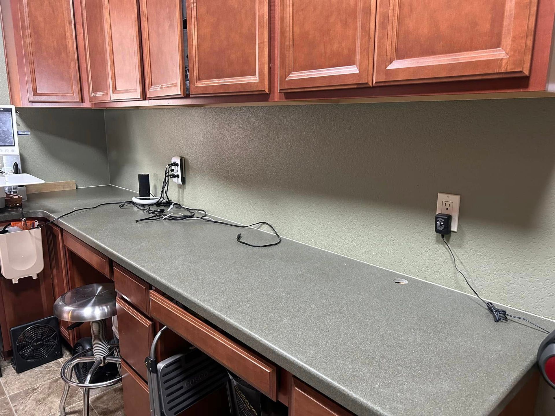 Long countertop under brown cabinets with cords, outlets, and a stool.