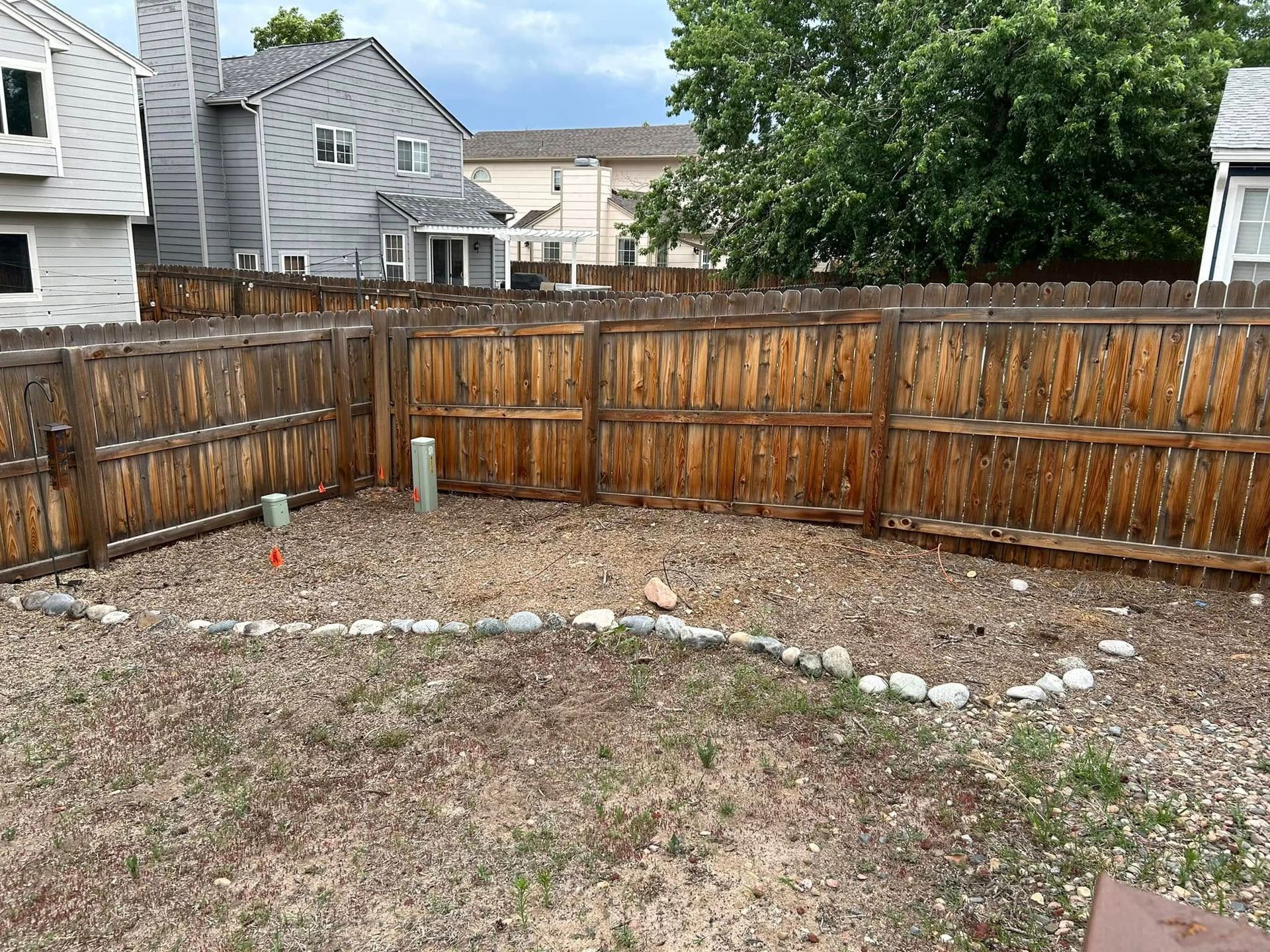 Backyard enclosed by a wooden fence with gravel ground and a rock border. Houses are visible in the background.