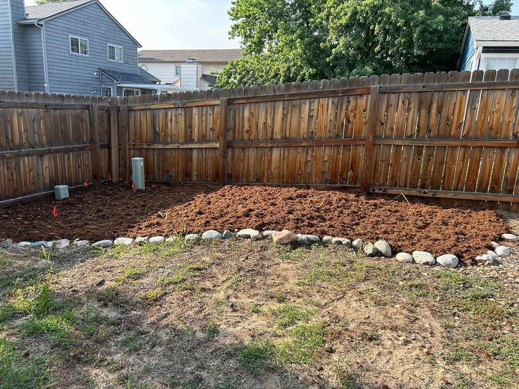 A backyard with a wood fence, mulch area, grass, and a few small rocks lining the perimeter.