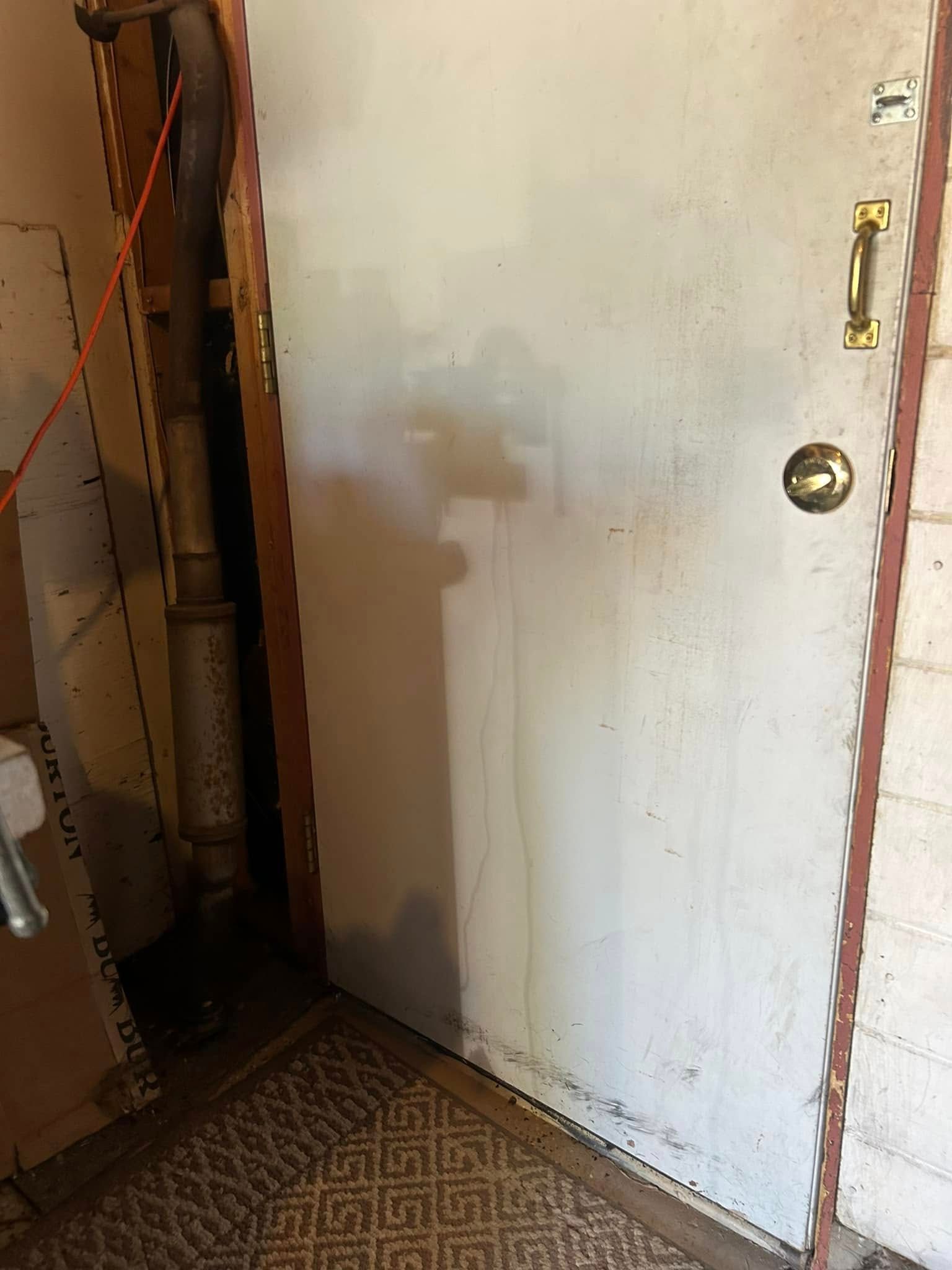 A dirty white door with brass hardware, next to a pipe and a brick wall.