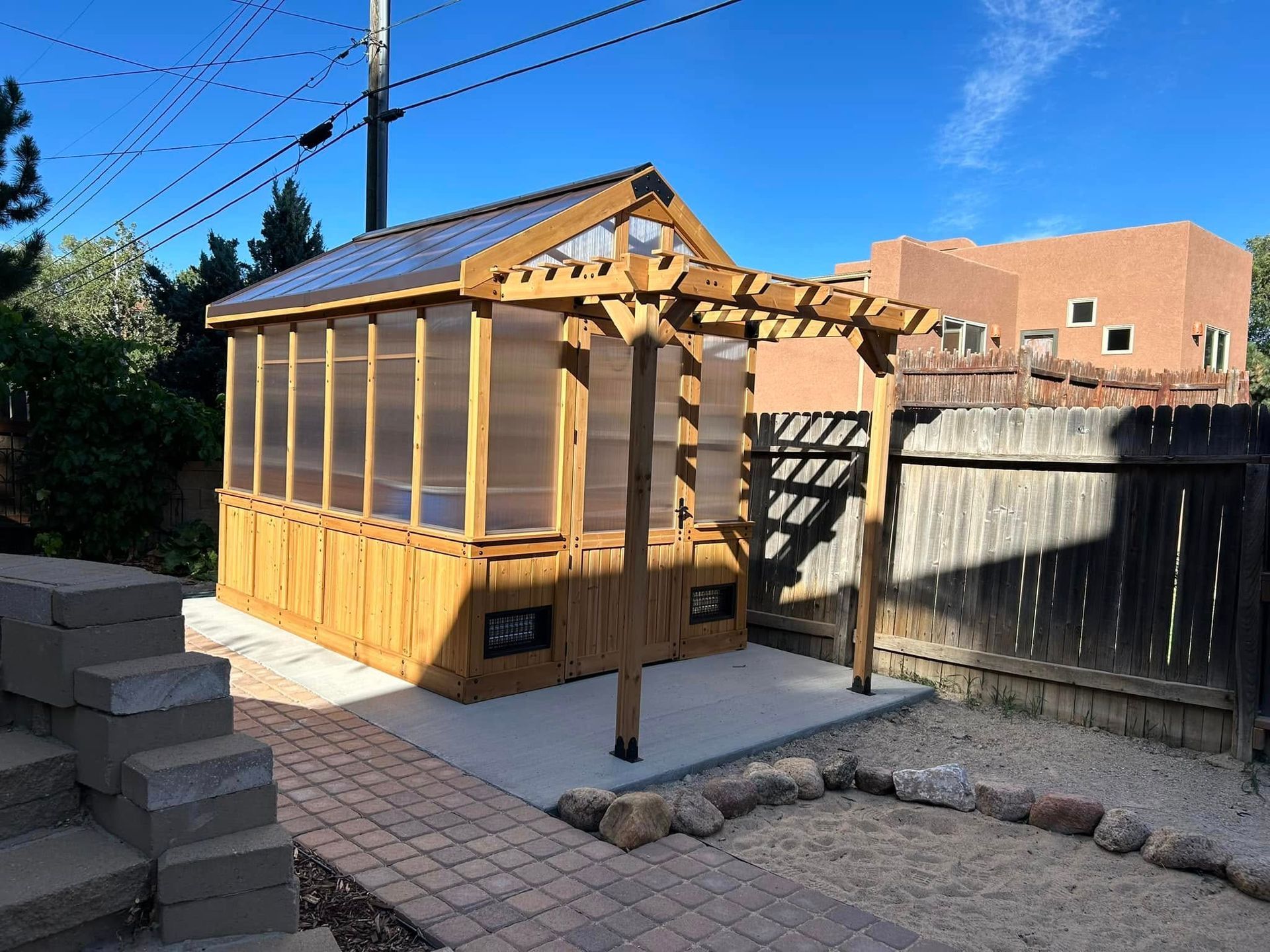 Wooden greenhouse with pergola in a sunny backyard, next to a brick patio.