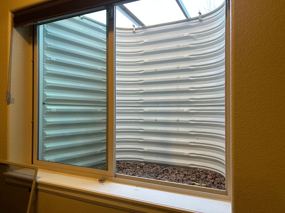 A sliding window reveals a corrugated window well with gravel, sunlight, and a clear view of the sky.