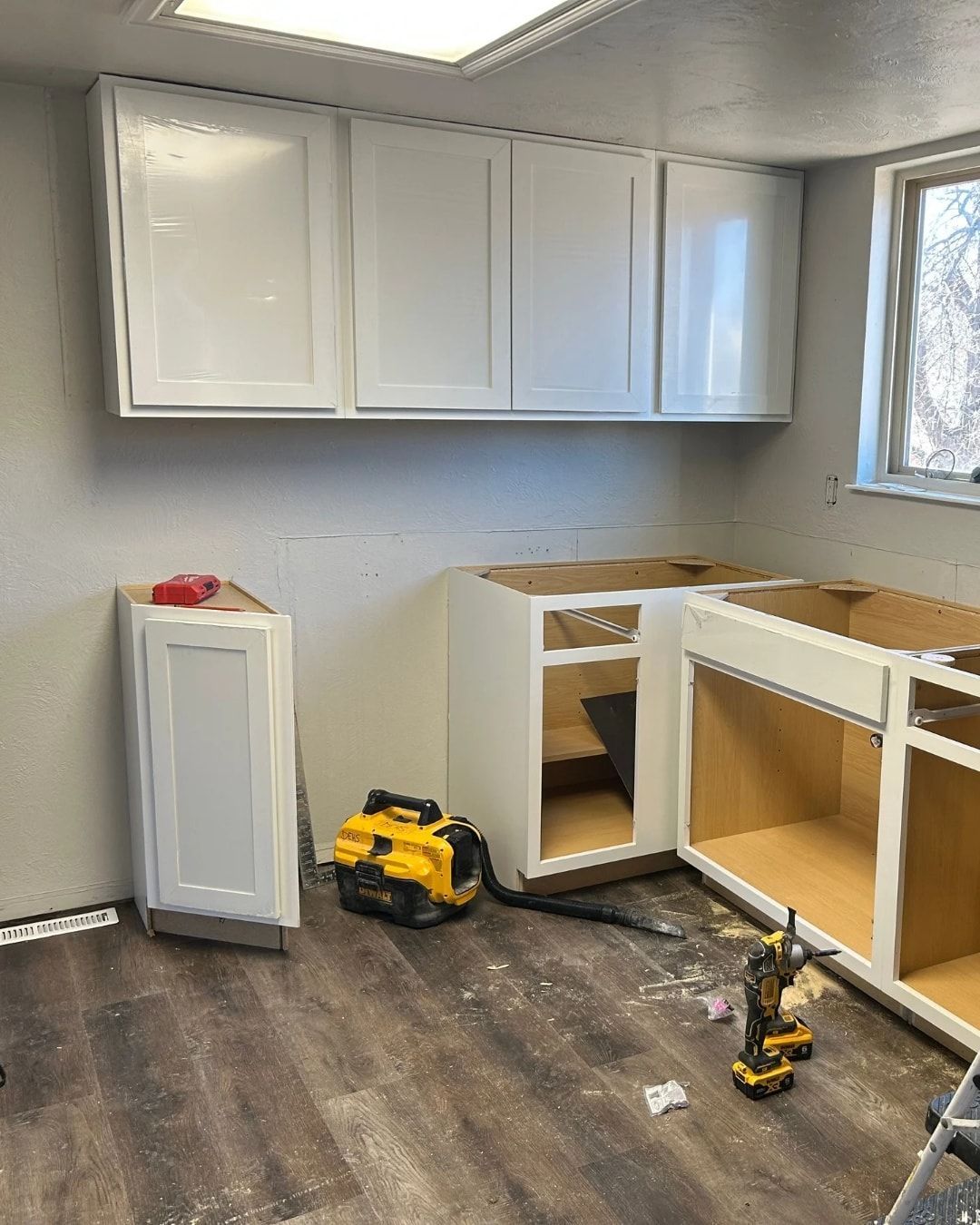 White kitchen cabinets being installed, with tools and unfinished surfaces on a dark wood-look floor.