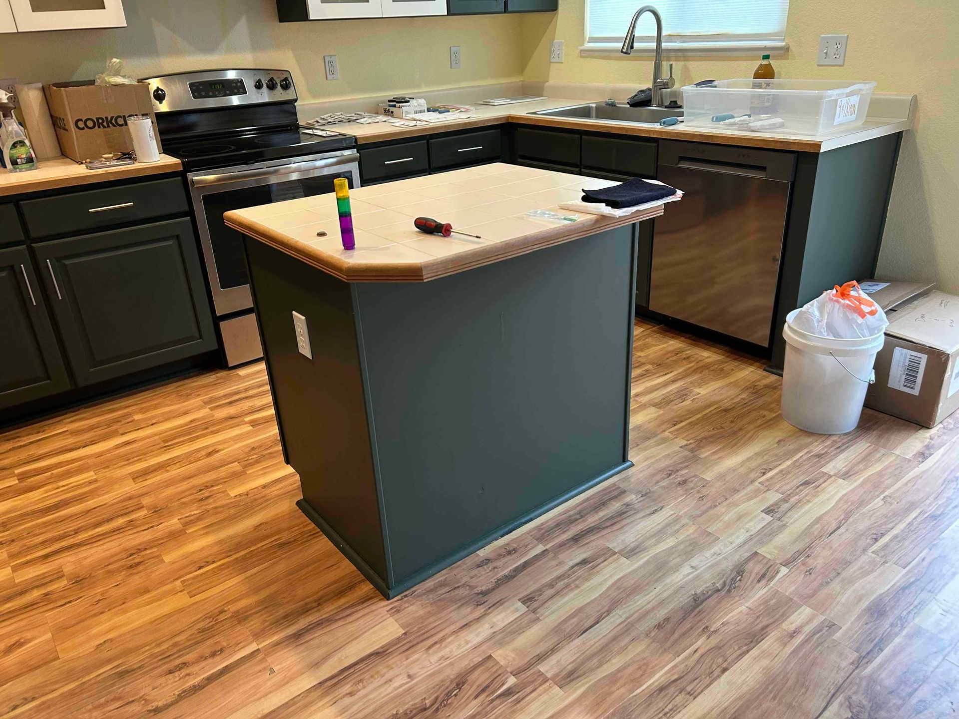 Kitchen with green cabinets, stainless steel appliances, and wood floors. An island with a light countertop is in the center.