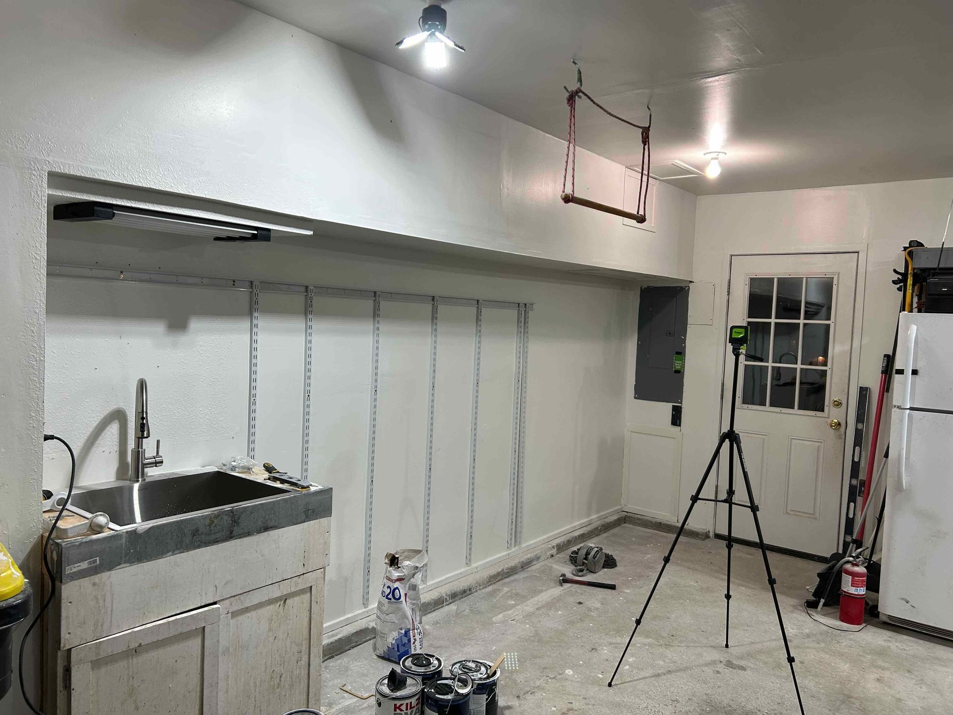 Garage interior with a utility sink, shelf supports, and a laser level on a tripod.