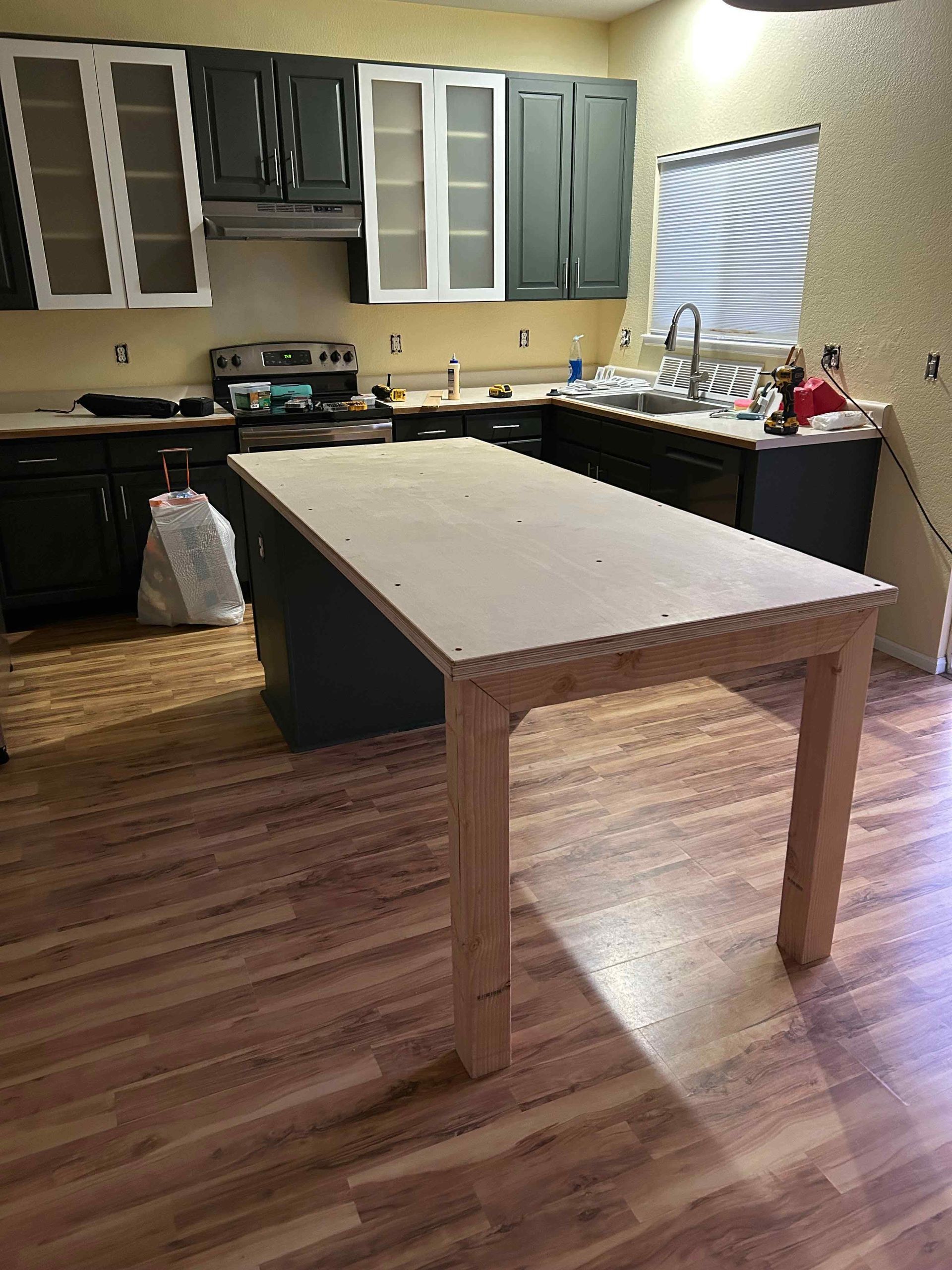 Kitchen undergoing renovation with an island and dark cabinets, unfinished wooden table, and wood-look flooring.