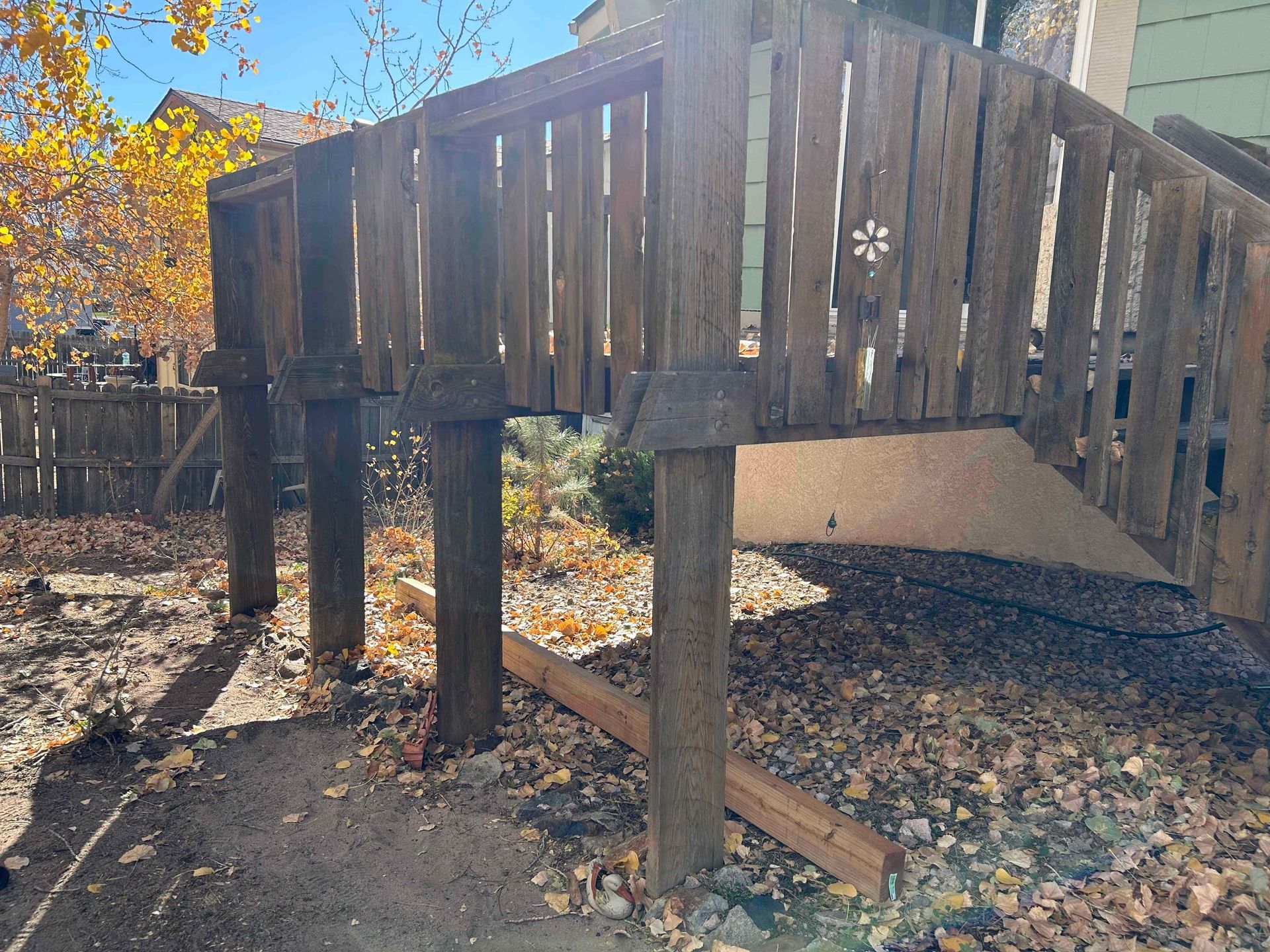 Wooden play structure with vertical supports, set in a yard with gravel and leaves.