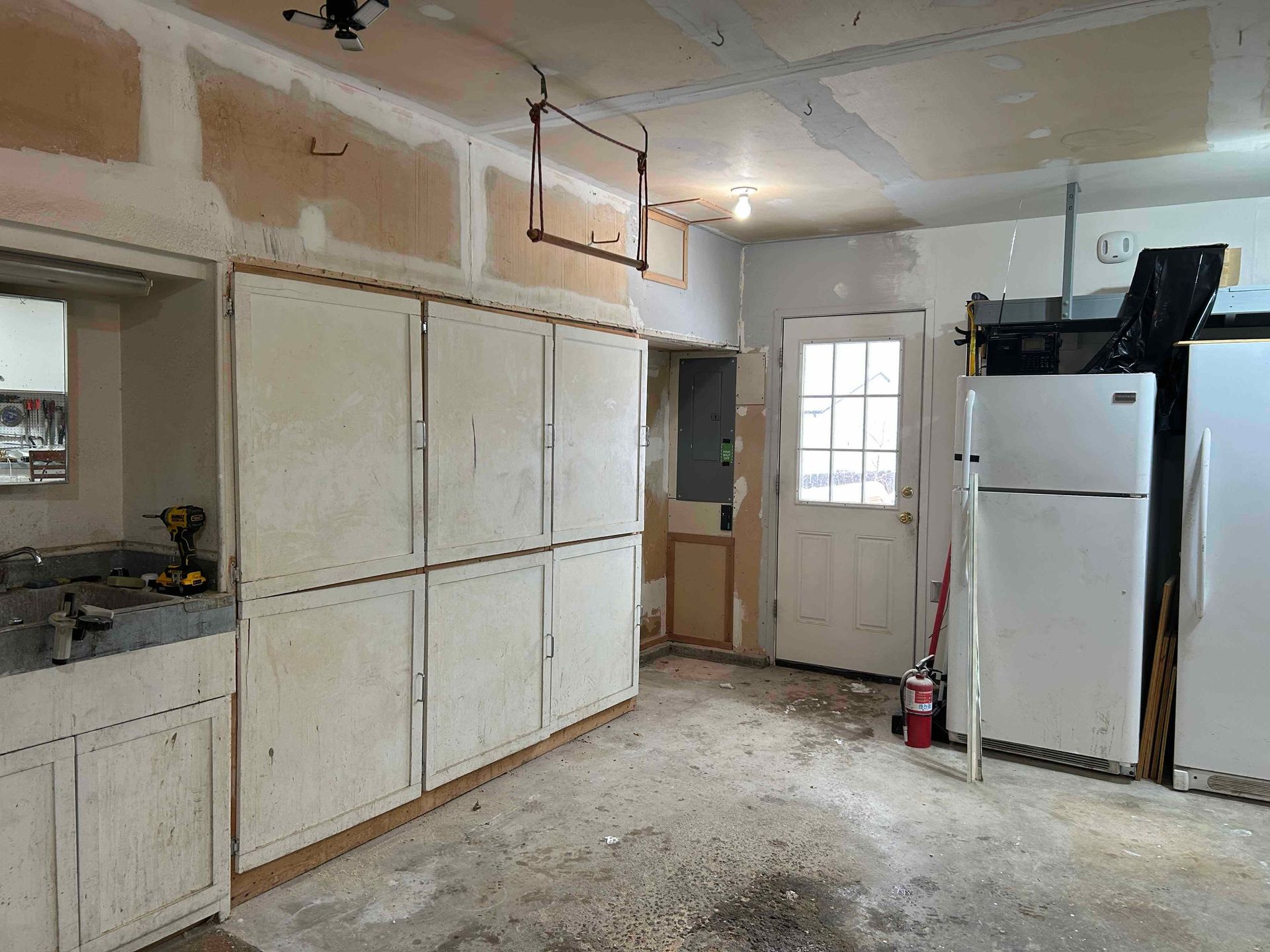 Garage interior with cabinets, door, appliances, and exposed wall. Concrete floor, damaged walls, overhead track.