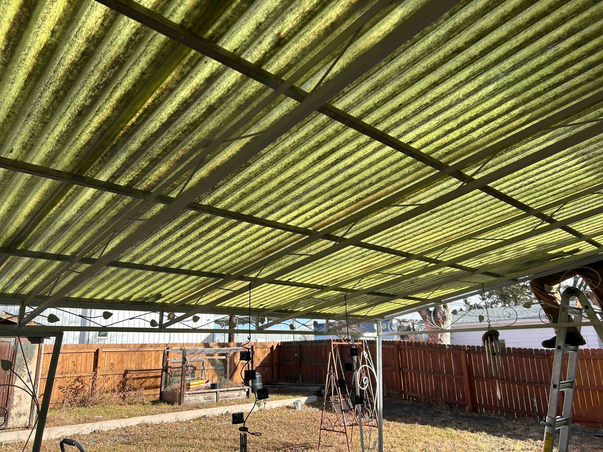 Green corrugated roof on a metal frame, covering a backyard with a fence and decorations.