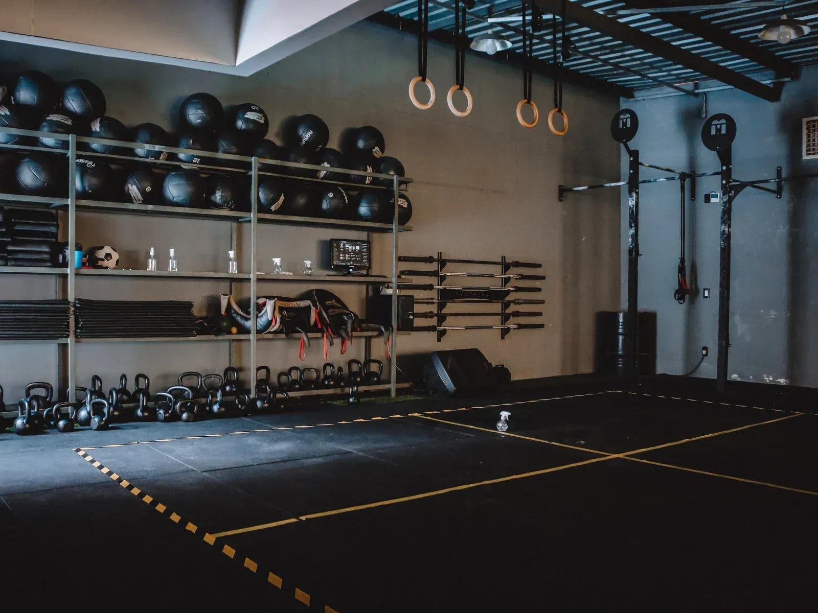 Gym interior with equipment: weights, rings, shelves of gear, and marked floor.