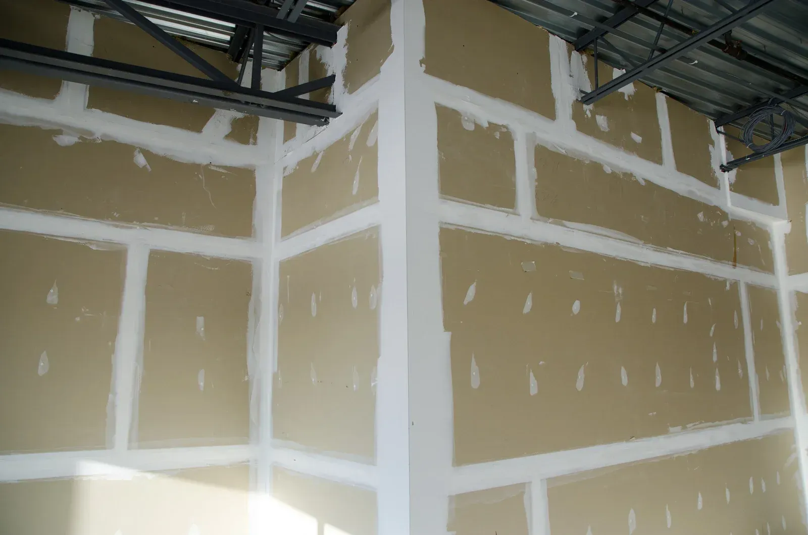 Man on a scaffold applying drywall compound to a ceiling with a trowel, near windows.