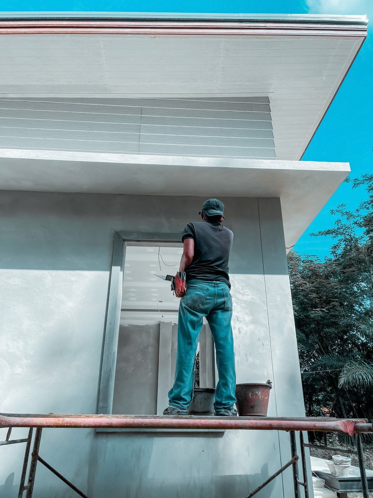 Construction worker on scaffolding drills into a light-colored building exterior; blue sky.