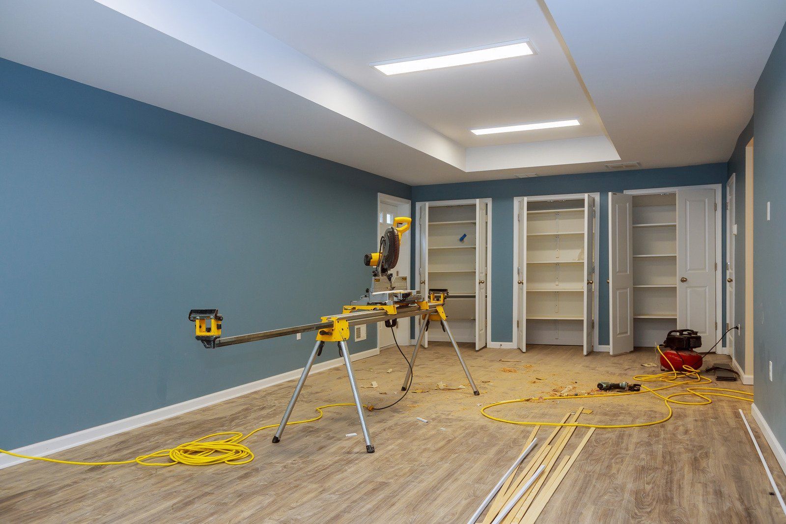 Construction in a room with blue walls, light wood-look flooring, and white cabinets. A miter saw sits on a stand.