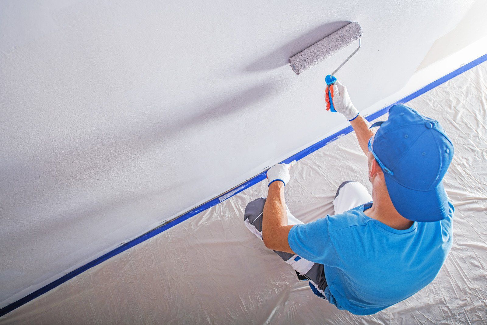 Person in blue painting a white ceiling with a roller; protective tape lines the edges, tarp covers the floor.