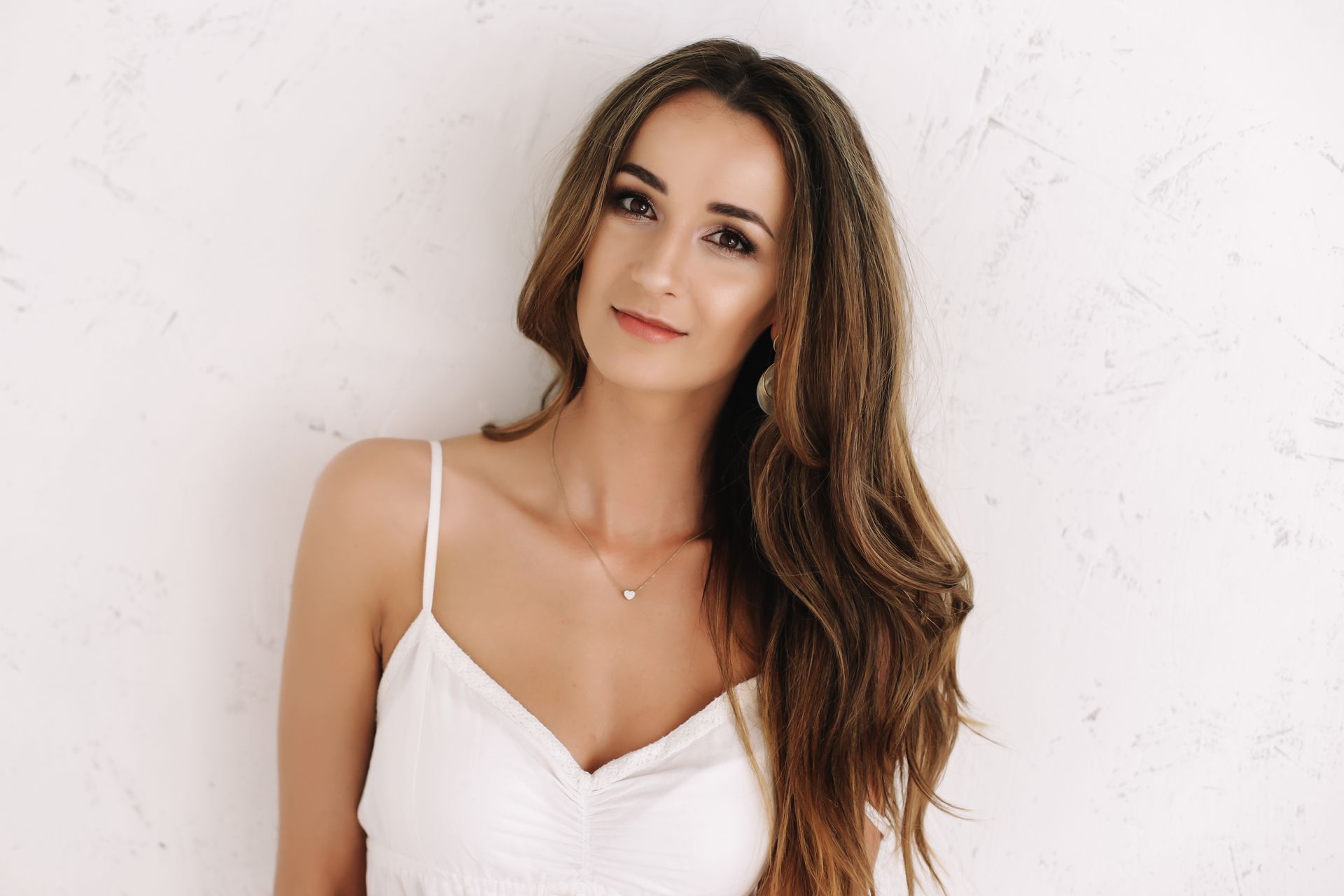 Woman with long brown hair, wearing a white dress, smiles in front of a white textured wall.