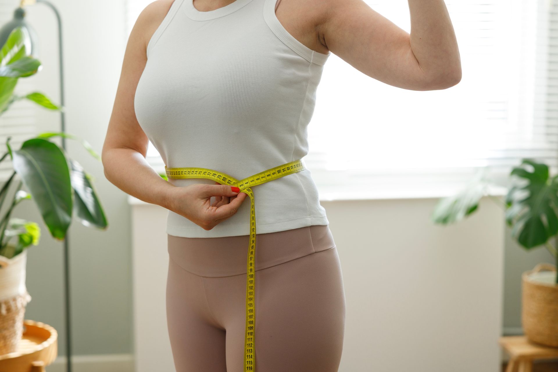 Woman in white tank top and leggings measuring her waist with yellow tape.