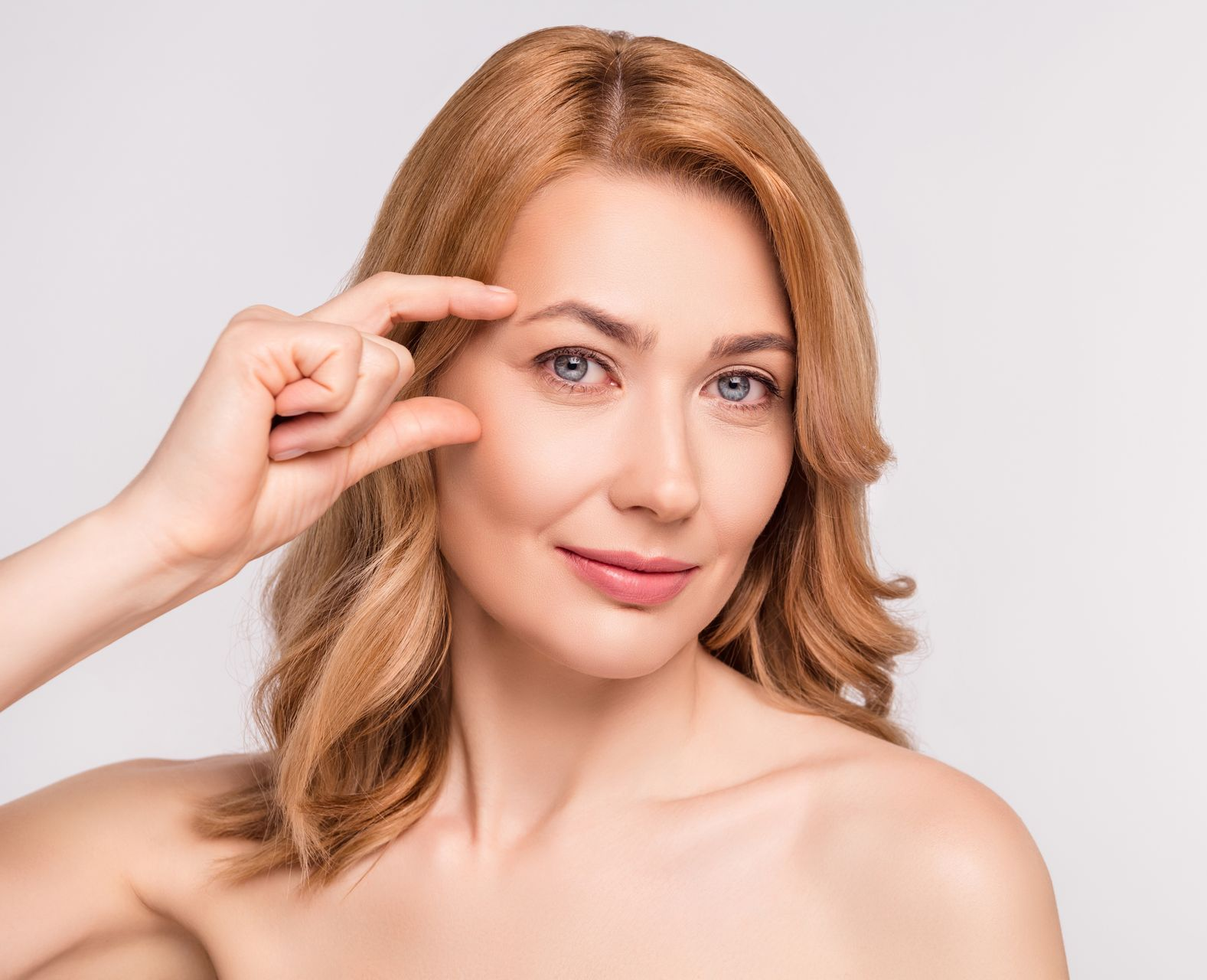 Woman with fair skin touches eyebrow, smiling. Light background.