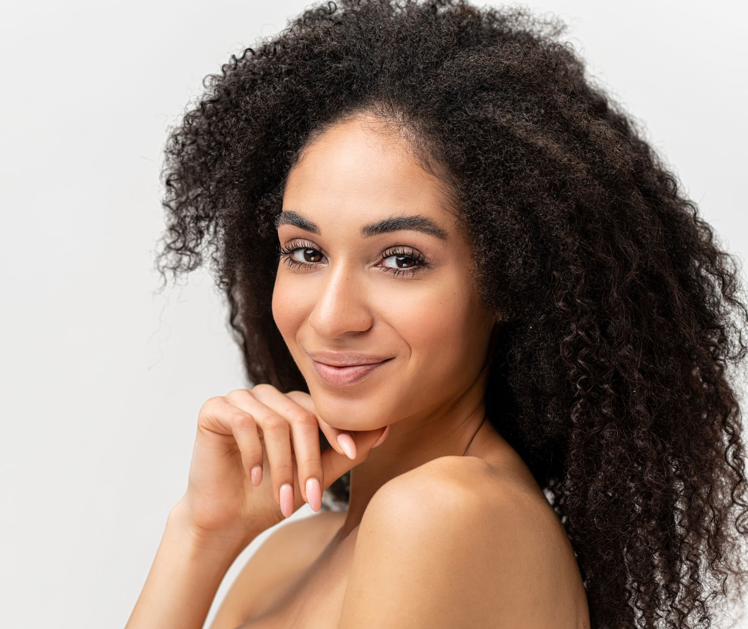 Woman with voluminous dark curly hair smiles, hand on chin, against a light background.