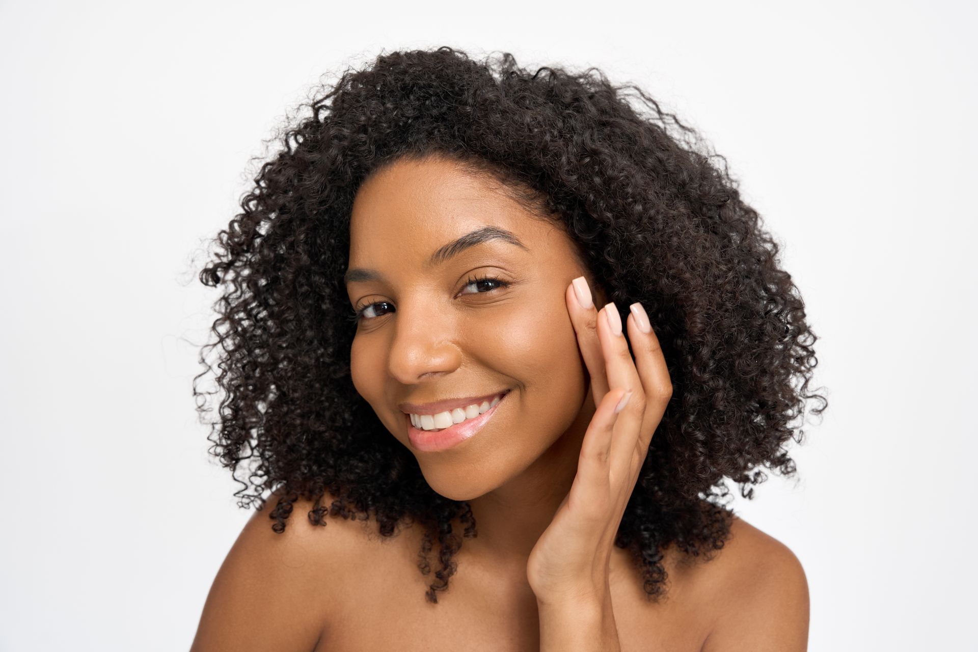A person with curly hair smiling while gently touching their cheek against a plain white background.