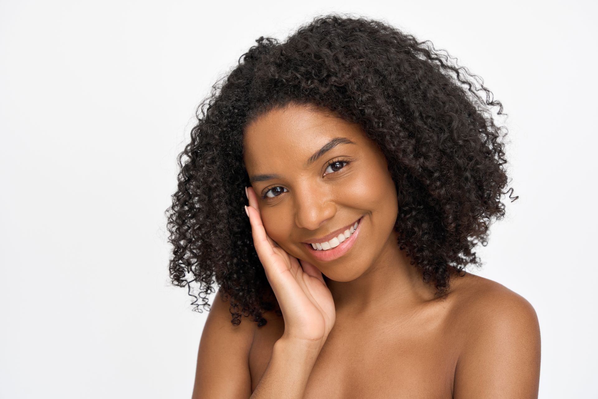 Woman with curly black hair smiles, touching face; bare shoulders, white background.